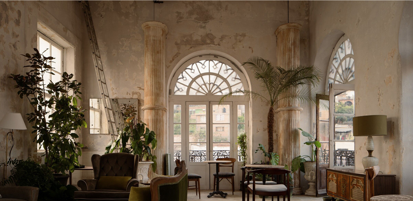 Interior de una sala con paredes desgastadas, columnas de estilo clásico, grandes ventanas con arcos y puertas de vidrio, mobiliario vintage y plantas decorativas.