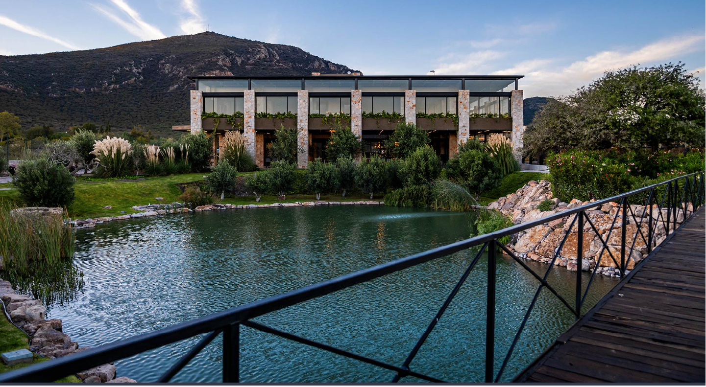 Edificio moderno con paredes de piedra y grandes ventanales, rodeado de vegetación y un estanque con un puente de madera en primer plano, montañas al fondo.