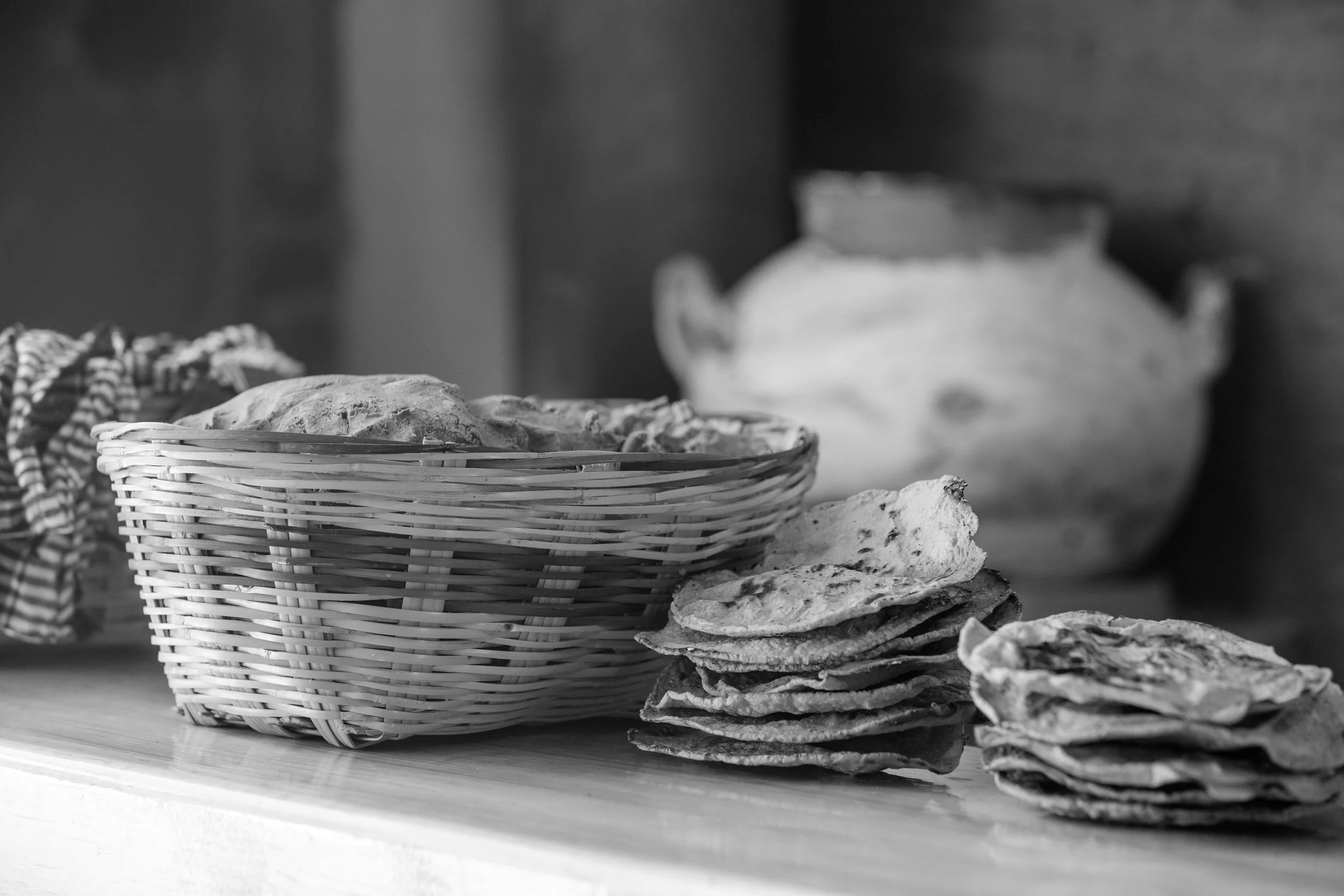 Montón de tortillas y pan en una cesta y sobre la mesa, con vasija en el fondo en un entorno rústico.