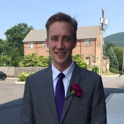 A young man in a gray suit with a purple tie and a pink flower pinned to his lapel, smiling outdoors near a car, with trees, power lines, and houses in the background.