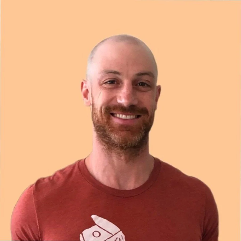 A smiling man with a beard and shaved head wearing a red t-shirt with a graphic design, standing against a peach-colored background.