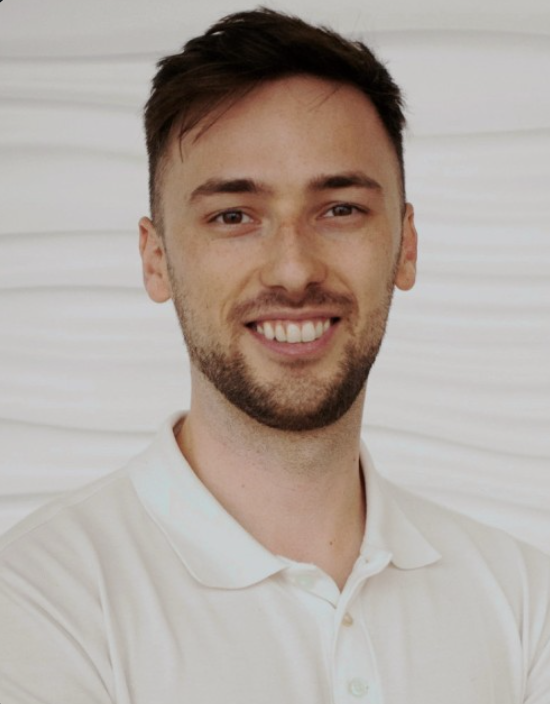 A smiling young man with brown hair and a beard, wearing a white polo shirt, standing in front of a white textured background.