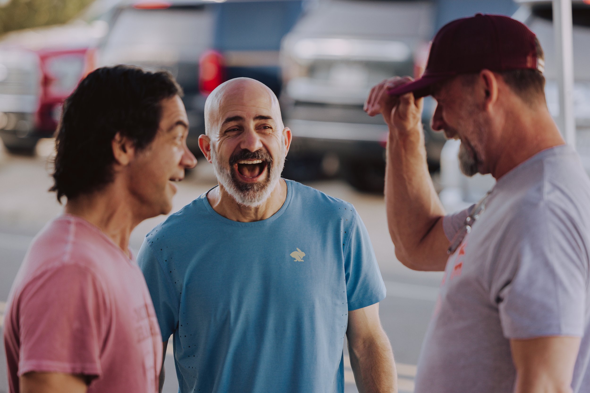 Three men smiling and talking outdoors near parked cars, one man is wearing a red cap and the others are in casual shirts.
