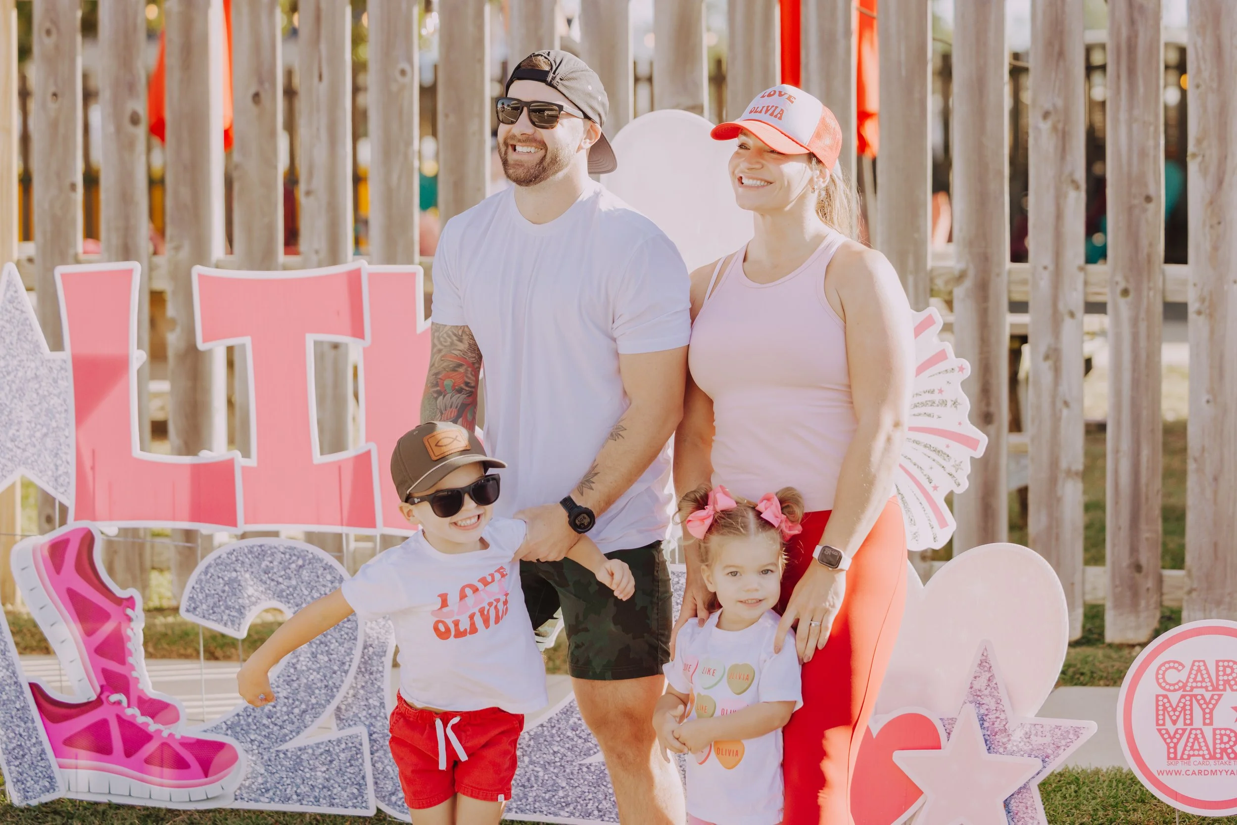 A smiling family of four, two adults and two children, standing outdoors at a fair or festival with decorative signs around them. The man has a beard, sunglasses, and a cap, while the woman wears a red cap and a light pink tank top. The children are wearing white shirts with red shorts and pink bows, and both kids are wearing sunglasses.