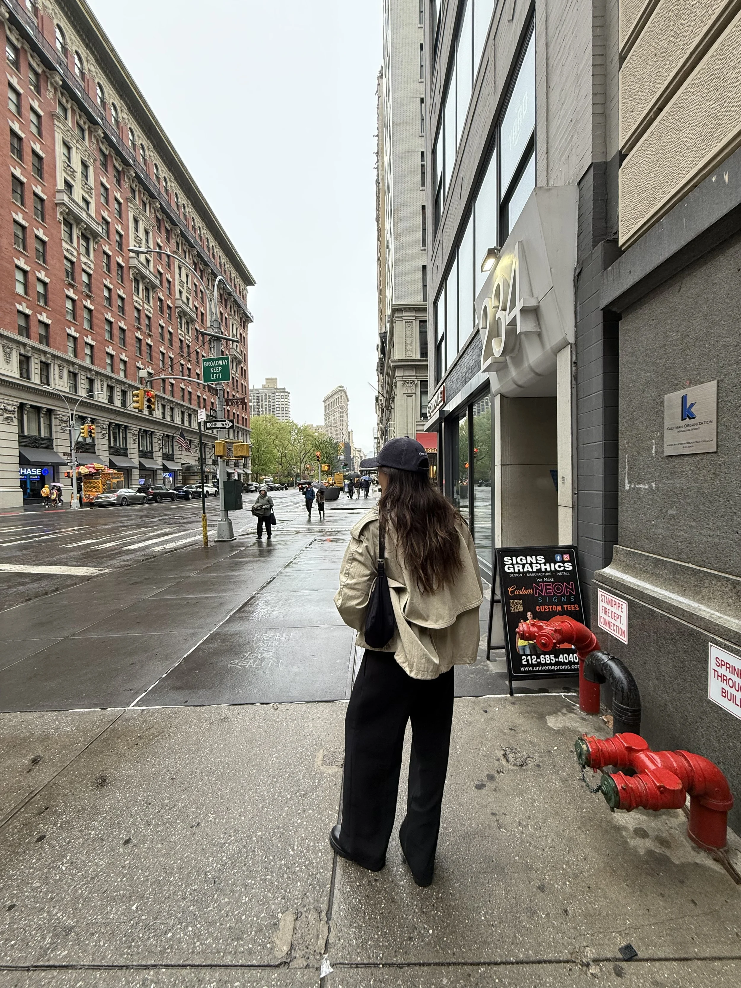 Woman standing on sidewalk wearing beige jacket and black wide-leg pants, holding walking cane, with buildings, street, and traffic in the background on a rainy day.