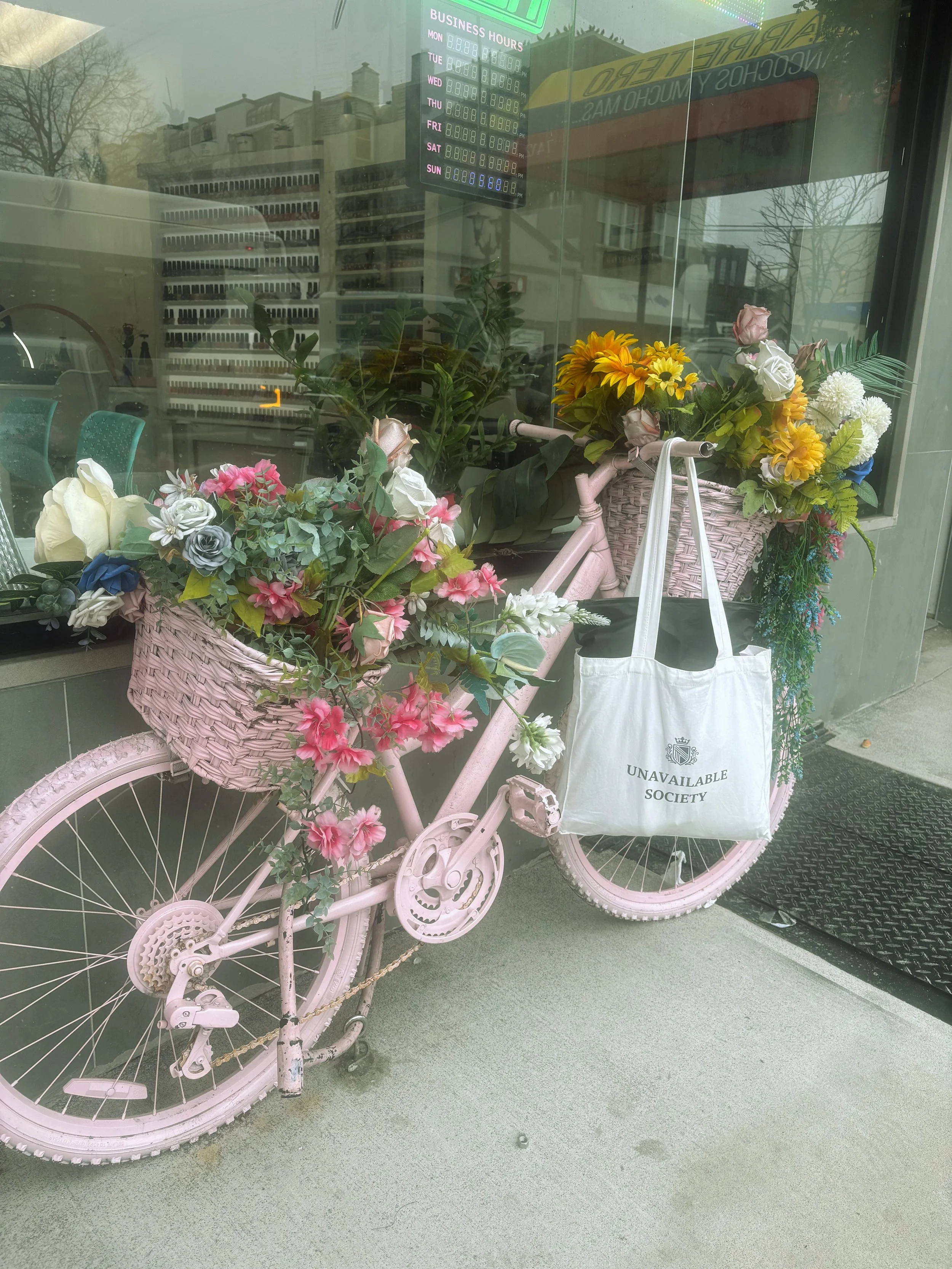 Pink bicycle with flower basket on the front, displayed in a store window. The basket contains colorful flowers, including roses and daisies. A white tote bag with the words "UNAVAILABLE SOCIETY" hangs from the bicycle's handlebar. Reflections of buildings and trees can be seen in the glass window.