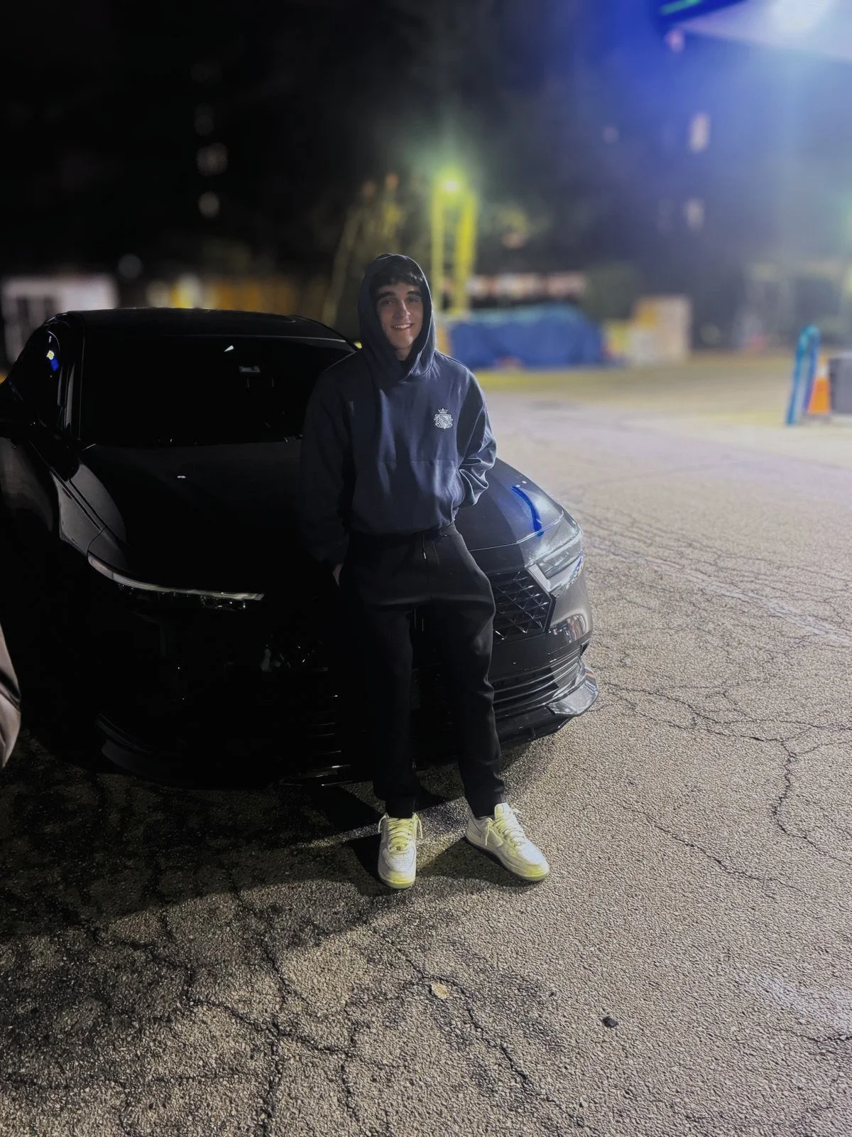A young man standing in front of a black car at night, wearing a blue hoodie, black pants, and yellow sneakers, with a parking lot and streetlights in the background.