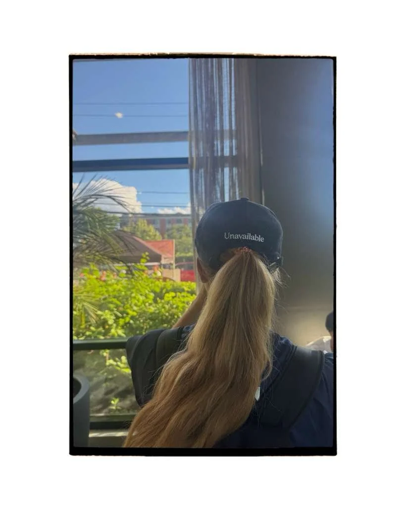 A person with long blonde hair wearing a black cap that says 'Unavailable' on it, sitting by a window with sheer curtains, looking outside on a sunny day with trees and buildings visible.