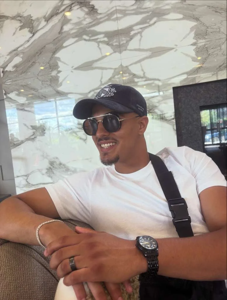A man sitting on a sofa in a modern indoor space, smiling, wearing sunglasses, a black baseball cap, a white t-shirt, a black watch, and a bracelet, with marble-clad walls and a television in the background.