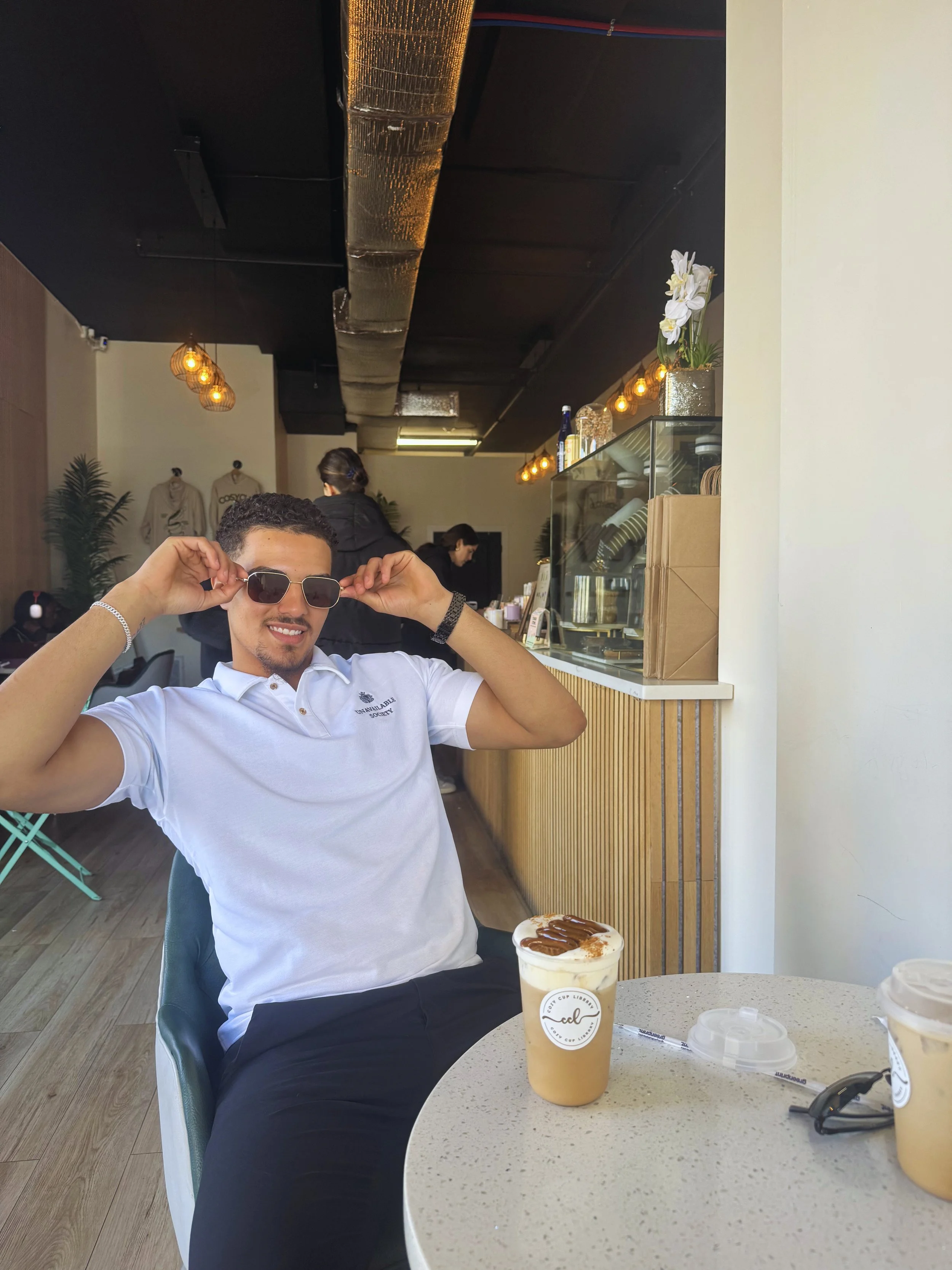 A man with short curly hair and sunglasses smiling and adjusting his sunglasses while sitting at a table in a café. There are two iced coffee drinks with chocolate syrup in front of him on the table. The café has modern decor with hanging lights, plants, and a display case.