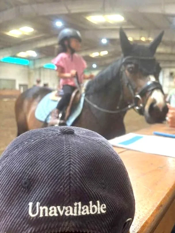 A baseball cap with the word "Unavailable" in front of a young girl riding a horse, wearing a helmet, in an indoor riding arena.
