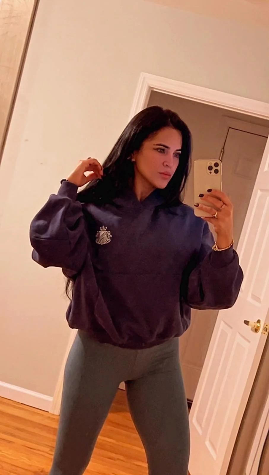 Woman taking a mirror selfie in her home, wearing a purple hoodie and gray pants.