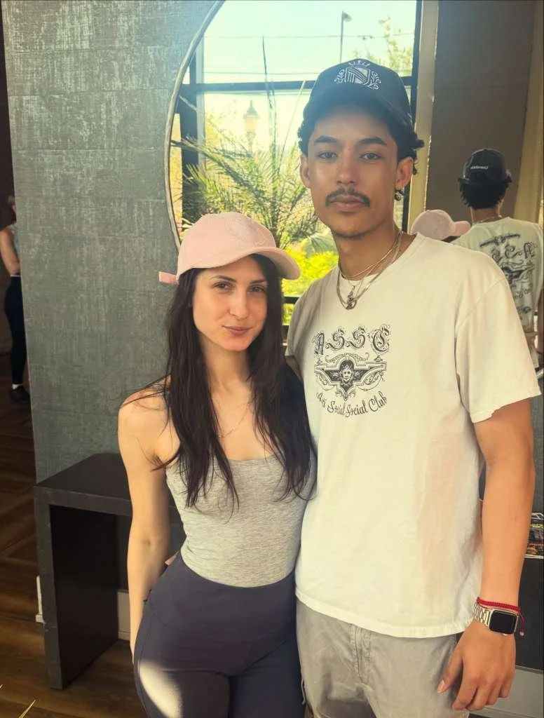 A young woman and a young man standing indoors, posing for a photo. The woman is wearing a light pink baseball cap, a gray tank top, and dark pants. The man is wearing a black cap, a white T-shirt with the number "1556" and emblems, and a watch on his left wrist. They are in a well-lit area with large windows showing trees and a building outside.