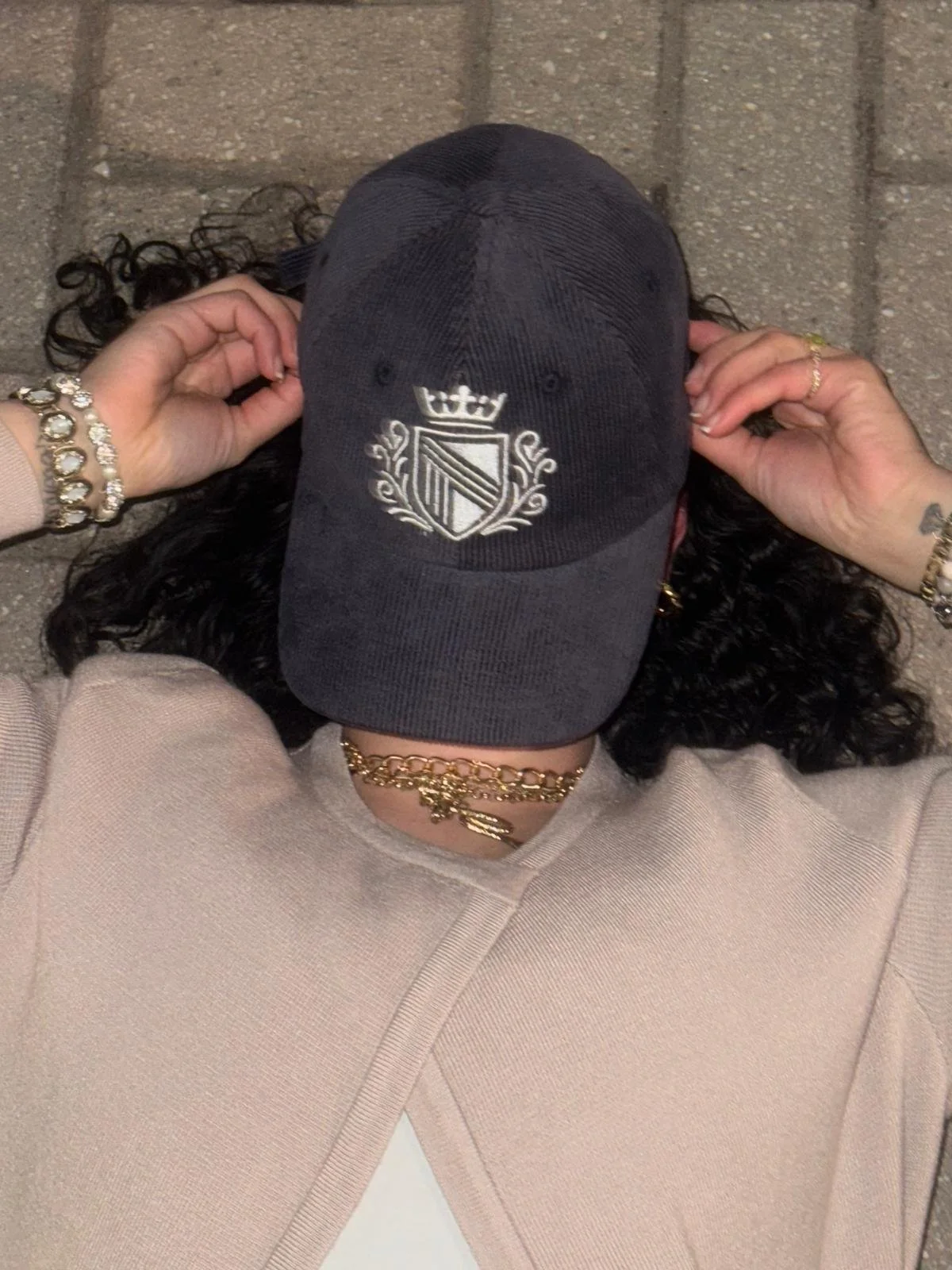 Person with curly dark hair, wearing multiple gold jewelry pieces, including bracelets, necklaces, and rings, holding a black cap with a shield and crown logo, lying on a pavement.