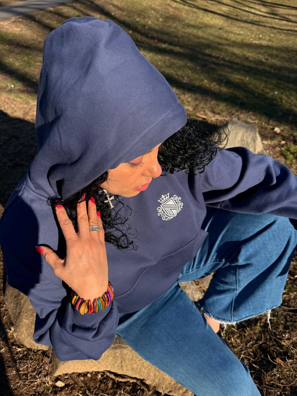 A woman with long curly black hair sitting outdoors on a rock, wearing a navy blue hoodie with a crest, blue jeans, with a colorful rainbow bracelet, cross earrings, and red nail polish, in a natural setting with sunlight and shadows.