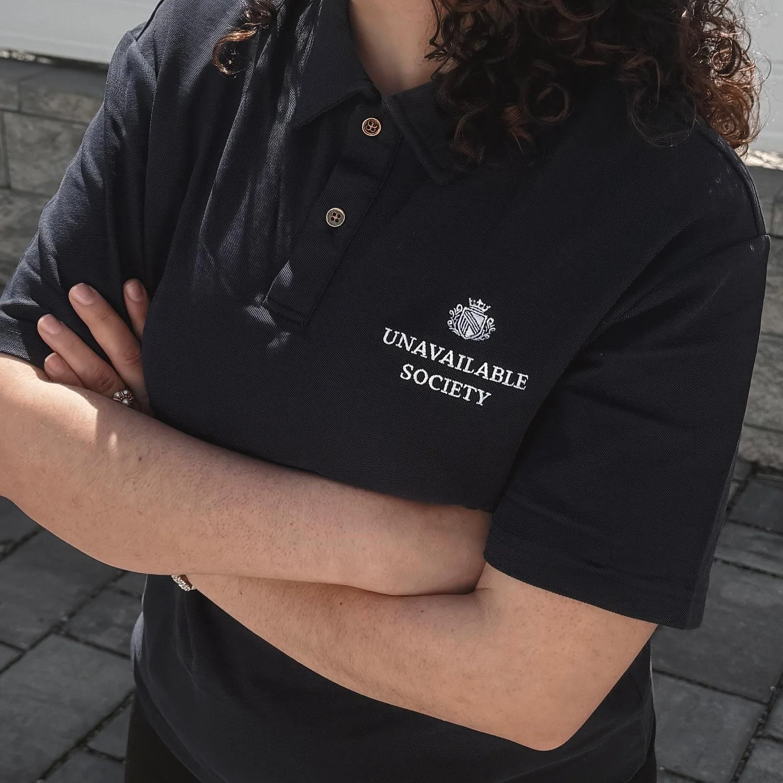A person with crossed arms wearing a black polo shirt with the logo and text 'Unavailable Society' on it, standing outdoors on a paved surface.