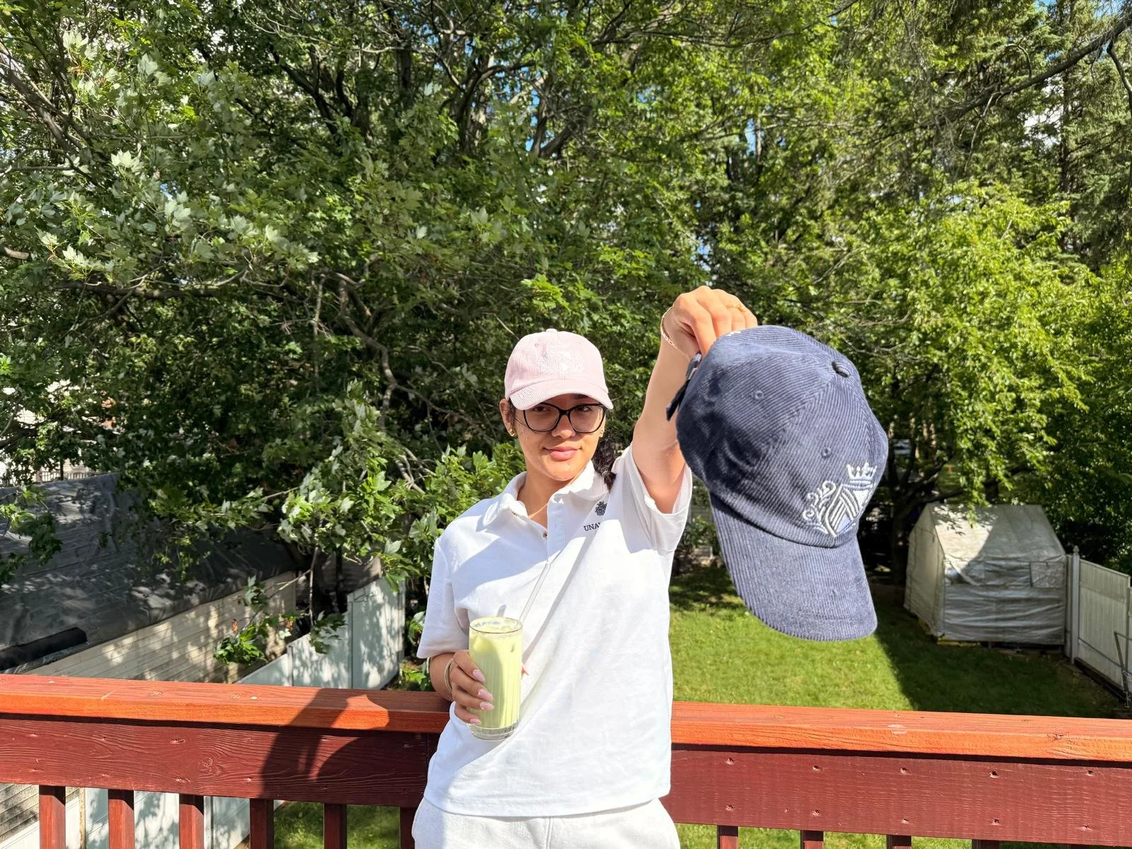 A woman wearing glasses and a light pink cap is standing on a wooden deck outdoors, holding a drink and swinging a blue baseball cap in the air. There are green trees and a white fence in the background.