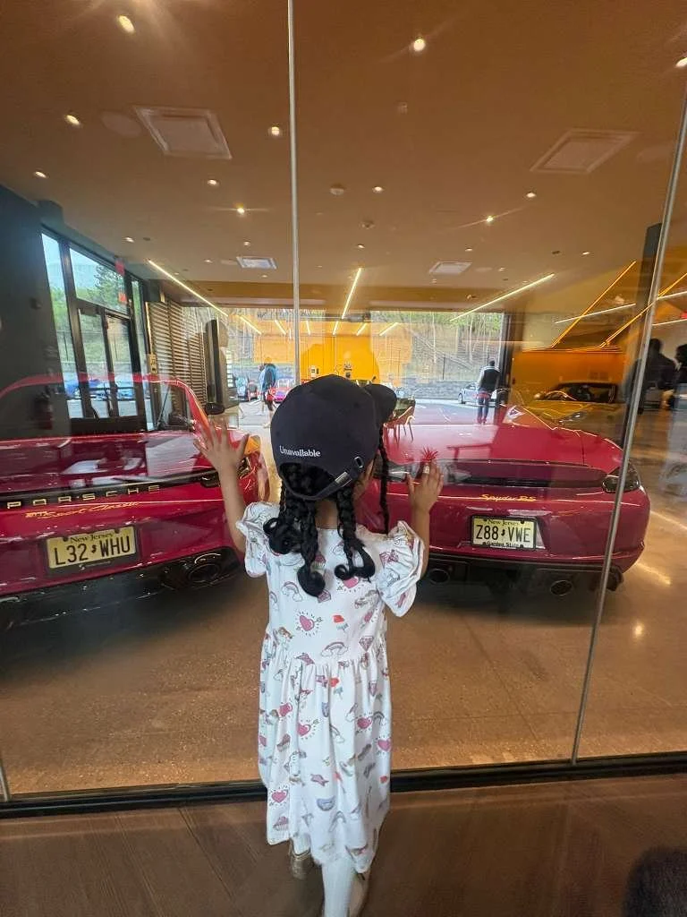 A young girl with black hair in pigtails, wearing a white dress with pink and blue flamingo patterns, stands behind a large glass window, looking at the red Porsche sports cars inside the showroom. She has raised hands pressed against the glass.