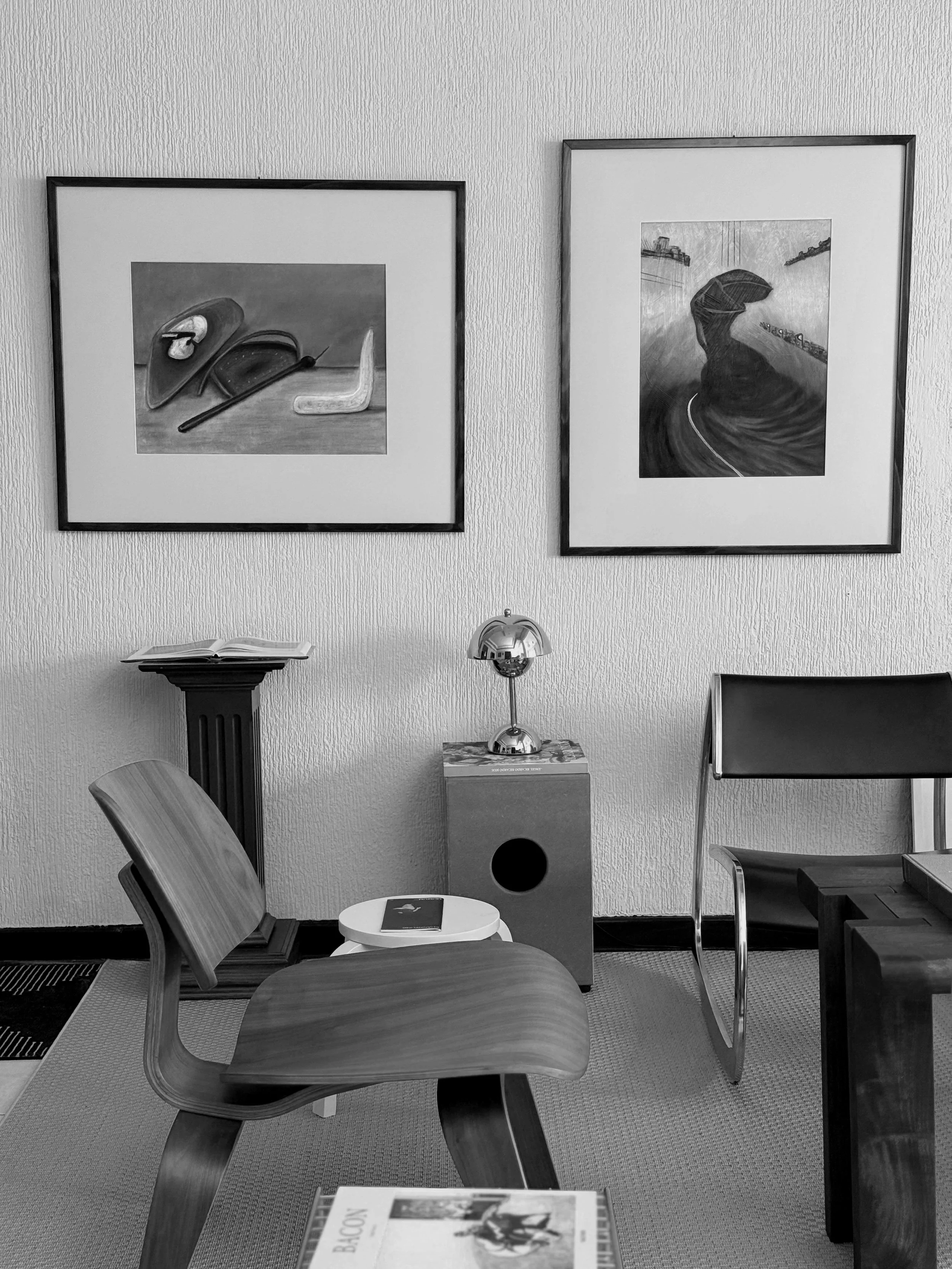 A black-and-white photo of an interior space featuring two framed artworks on the wall, a wooden chair, a table with a magazine, a small white side table, and a modern lamp on a pedestal.