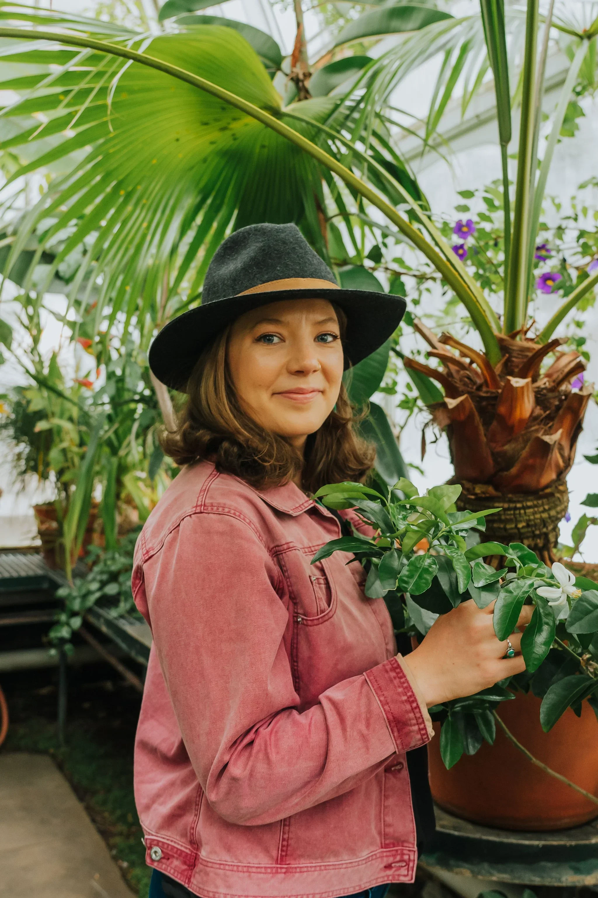 A woman in a pink jacket and dark gray hat standing among tropical plants and holding a small potted green plant.