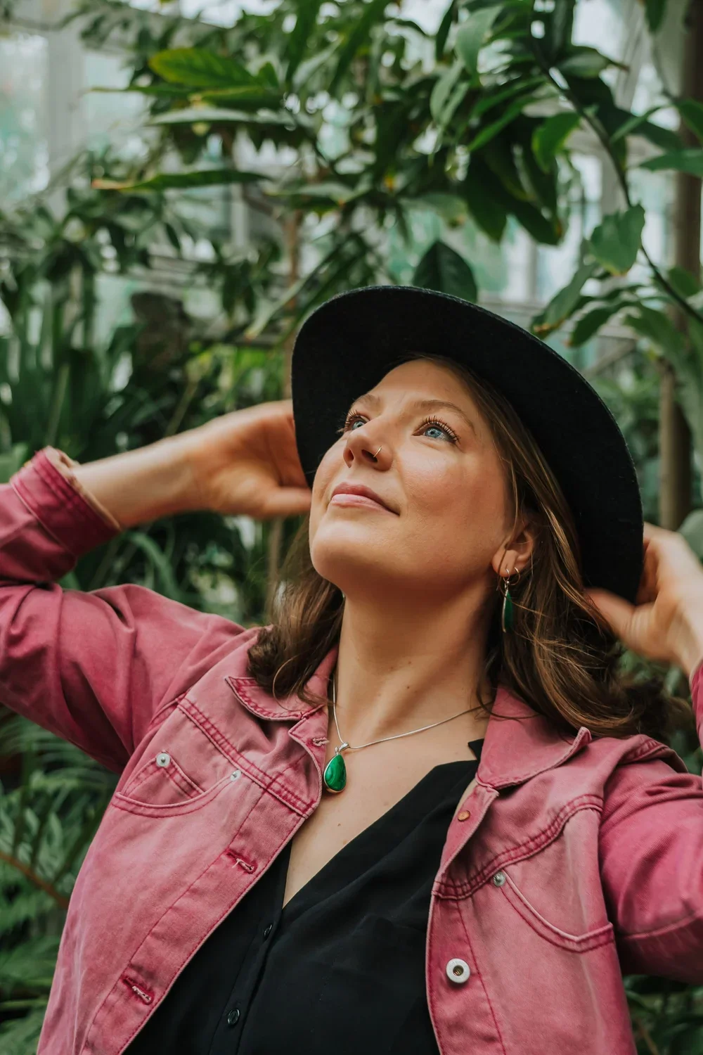 A woman wearing a black wide-brim hat, pink jacket, and green jewelry, smiling and looking upwards in a lush, green indoor garden.