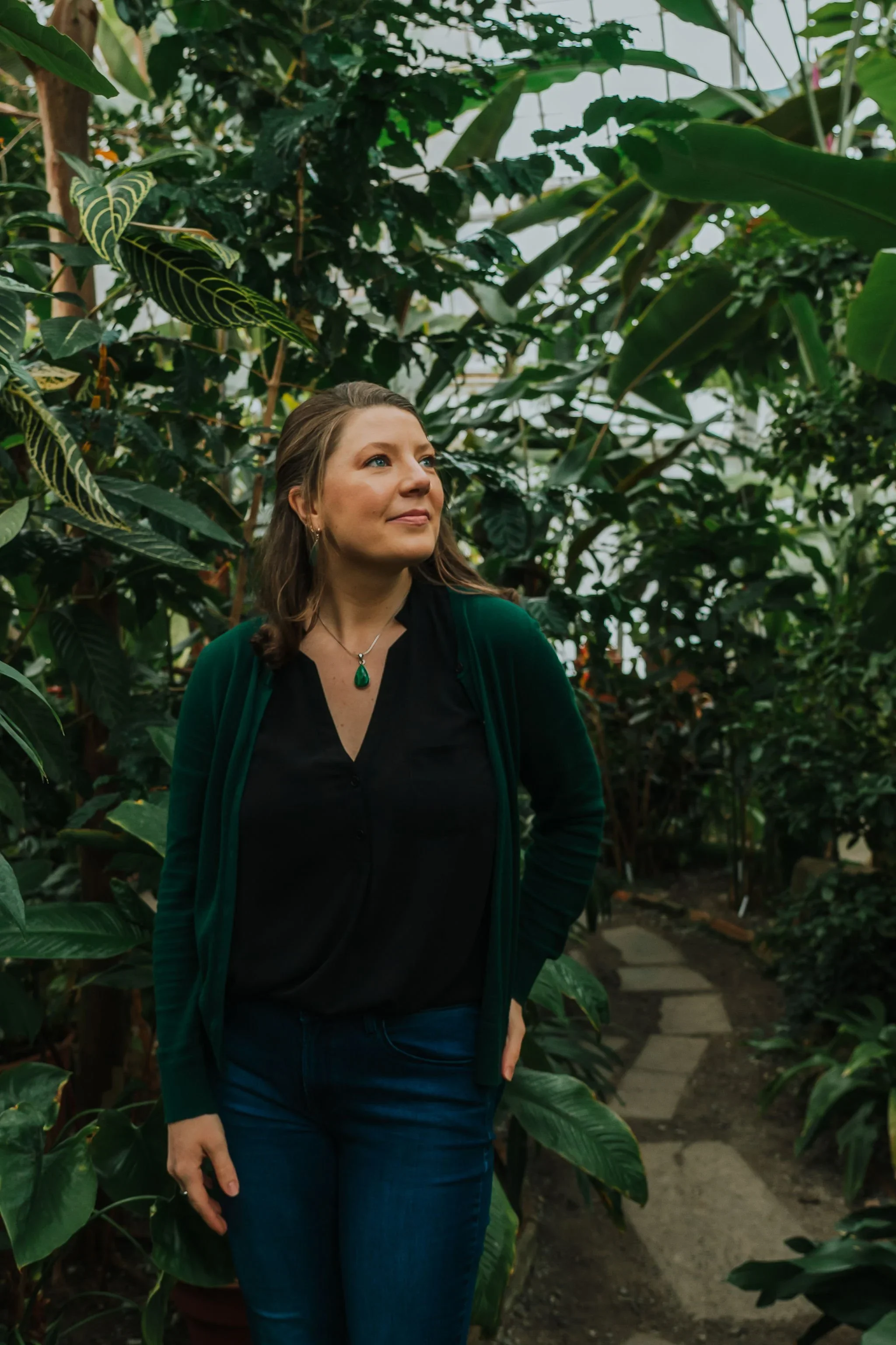 A woman standing in a lush, green botanical garden or greenhouse, surrounded by large tropical plants, wearing a black shirt, green cardigan, and blue jeans, looking off to the side.