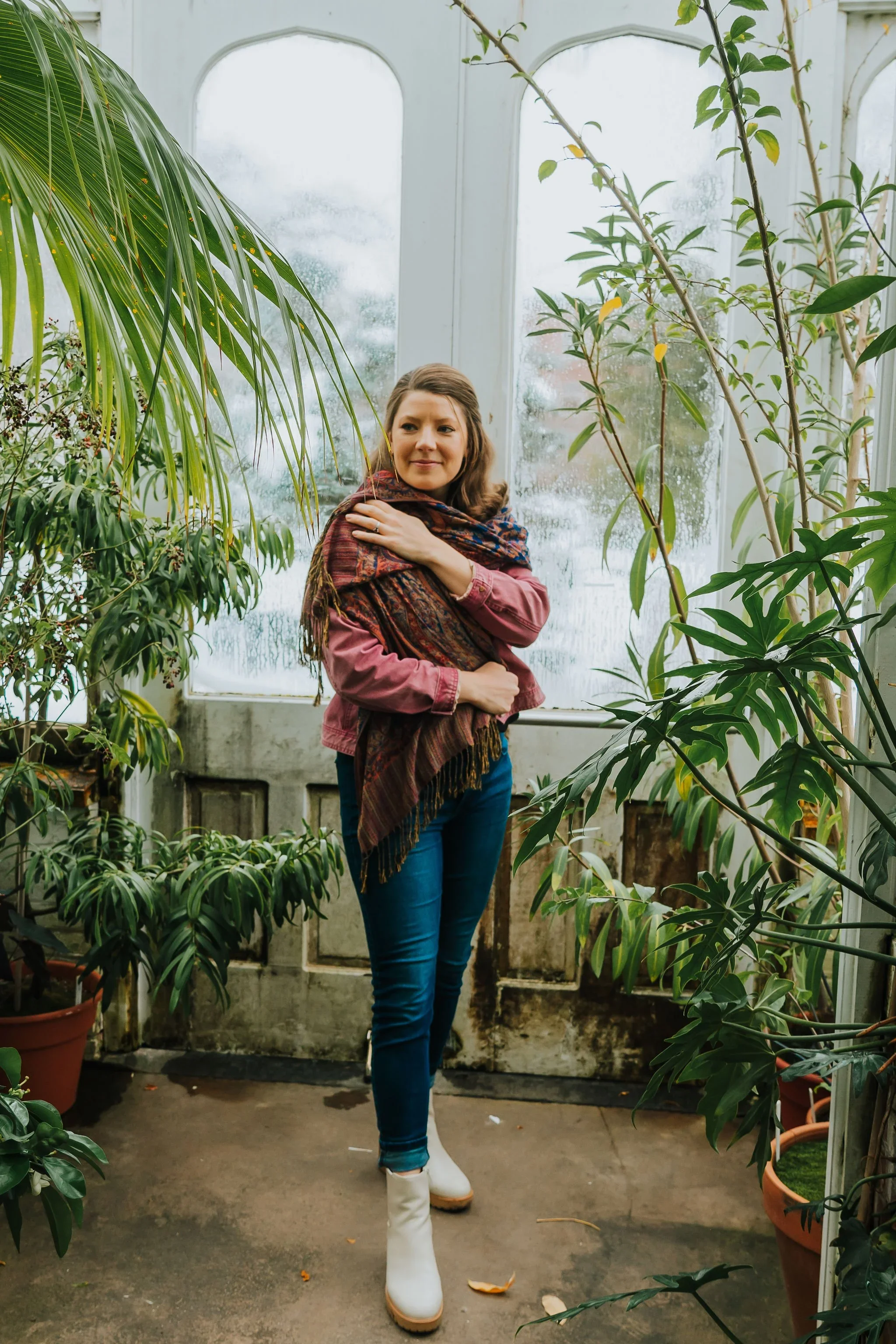 A woman in a greenhouse surrounded by various green plants. She is wearing a pink jacket, blue jeans, white boots, and a colorful scarf, and she is touching her shoulder while looking slightly to the side.