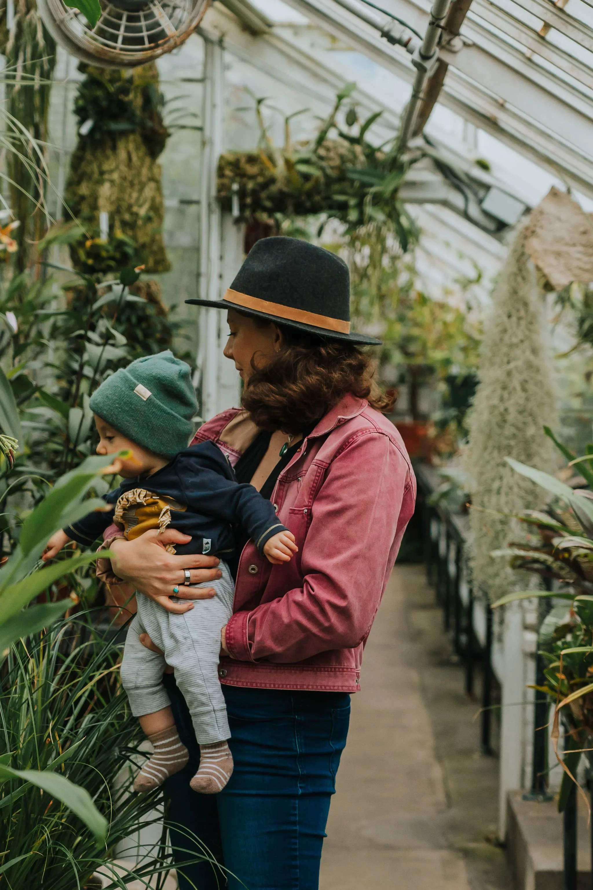 A woman in a pink jacket and a wide-brimmed hat holding a young child in a greenhouse, surrounded by tall plants and flowers.
