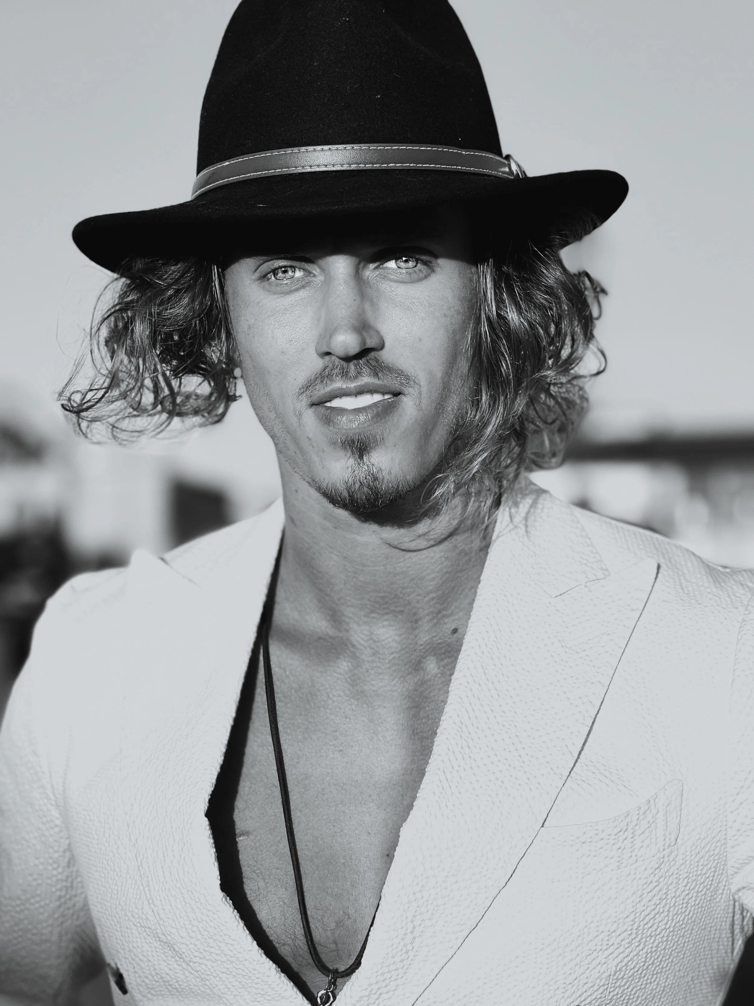 A man with long curly hair and a goatee, wearing a fedora hat and a light-colored textured blazer with an open collar, outdoors with a blurred background.