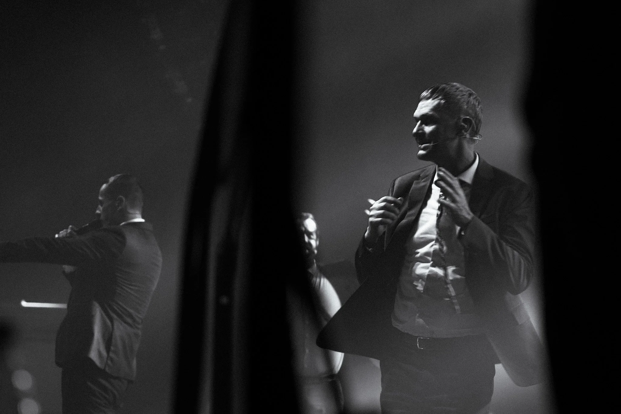 Black-and-white photo of a man in a suit speaking on stage, with two other people partially visible in the background, behind a curtain or partition.