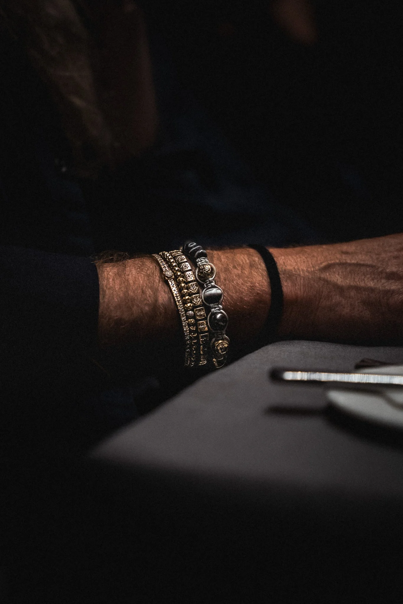 Close-up of a person's arm resting on a surface, wearing multiple black and gold bracelets. The background is dark and out of focus.