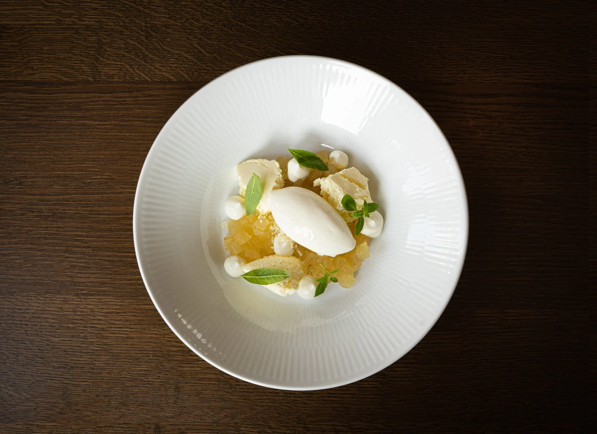 Dessert with a scoop of white ice cream, lemon custard, white chocolate pieces, small green leaves, and dollops of cream on a white plate