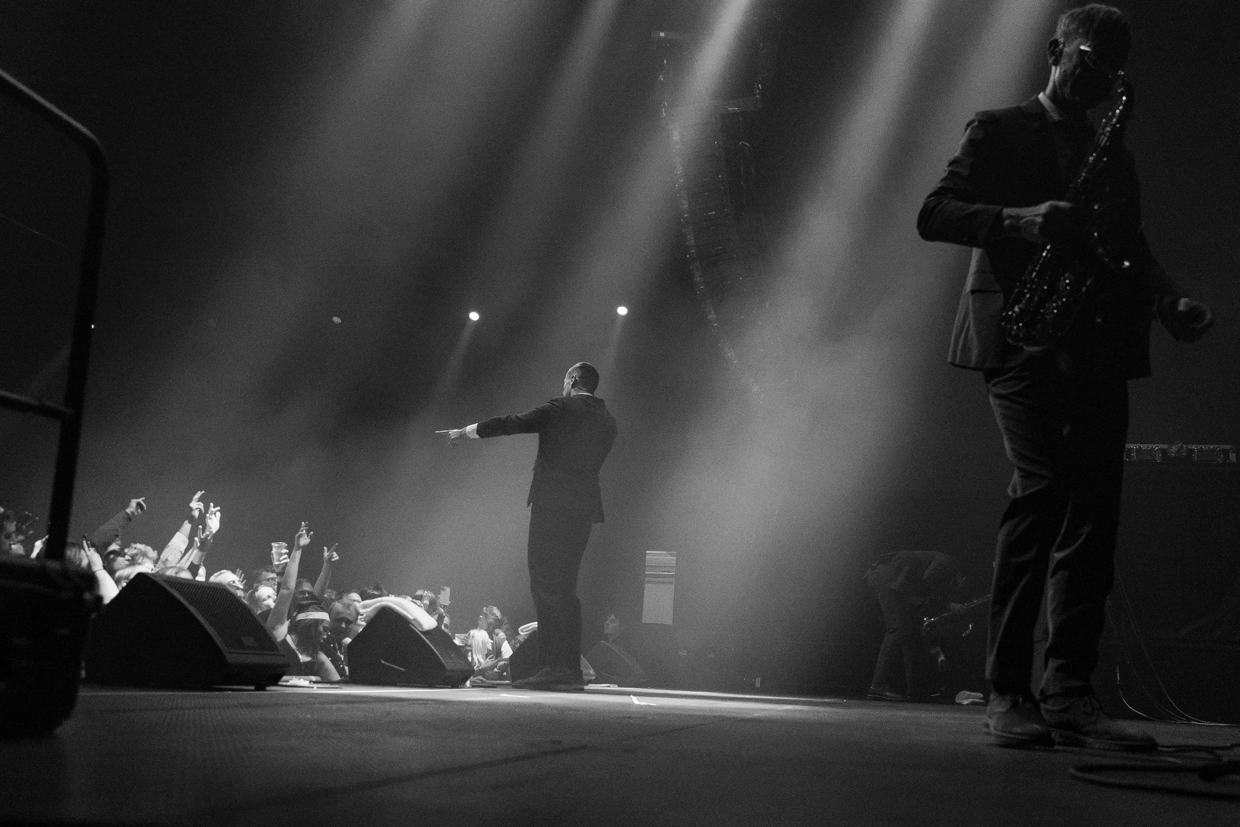 A black and white photo of a concert stage with a performer in a suit pointing to the audience, a saxophonist playing on the right, and a crowd with raised hands in front of the stage.