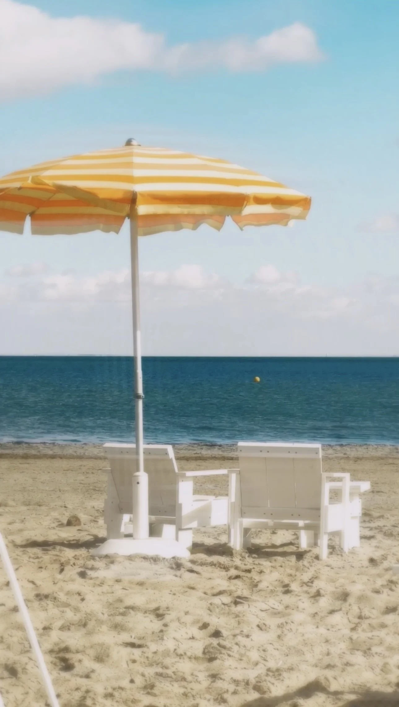 A beach scene with a yellow and white striped umbrella, two white deck chairs, and the ocean in the background under a partly cloudy sky.