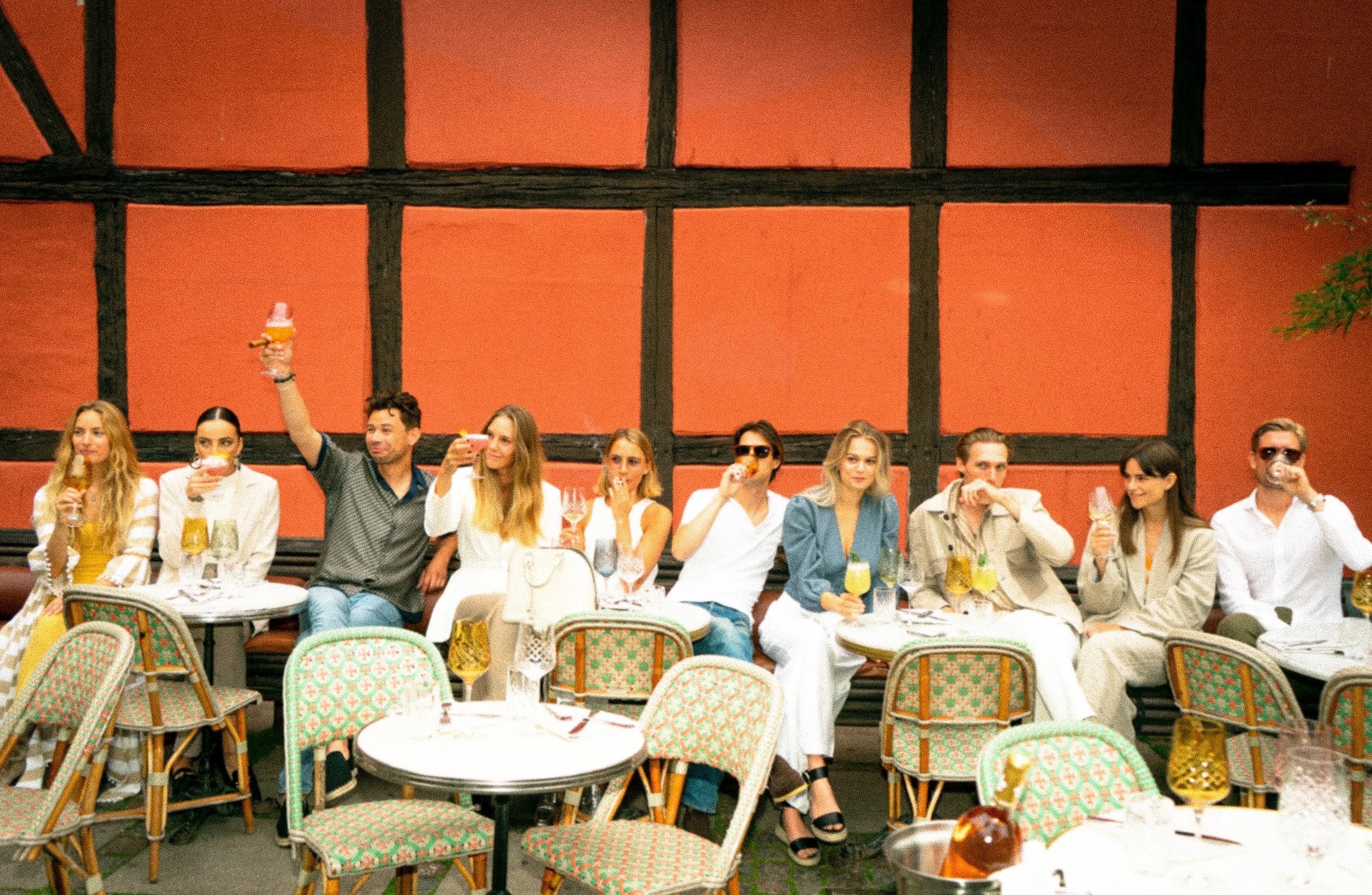 Group of people sitting at a long table raising glasses for a toast in a restaurant with a red wall and black wooden beams.