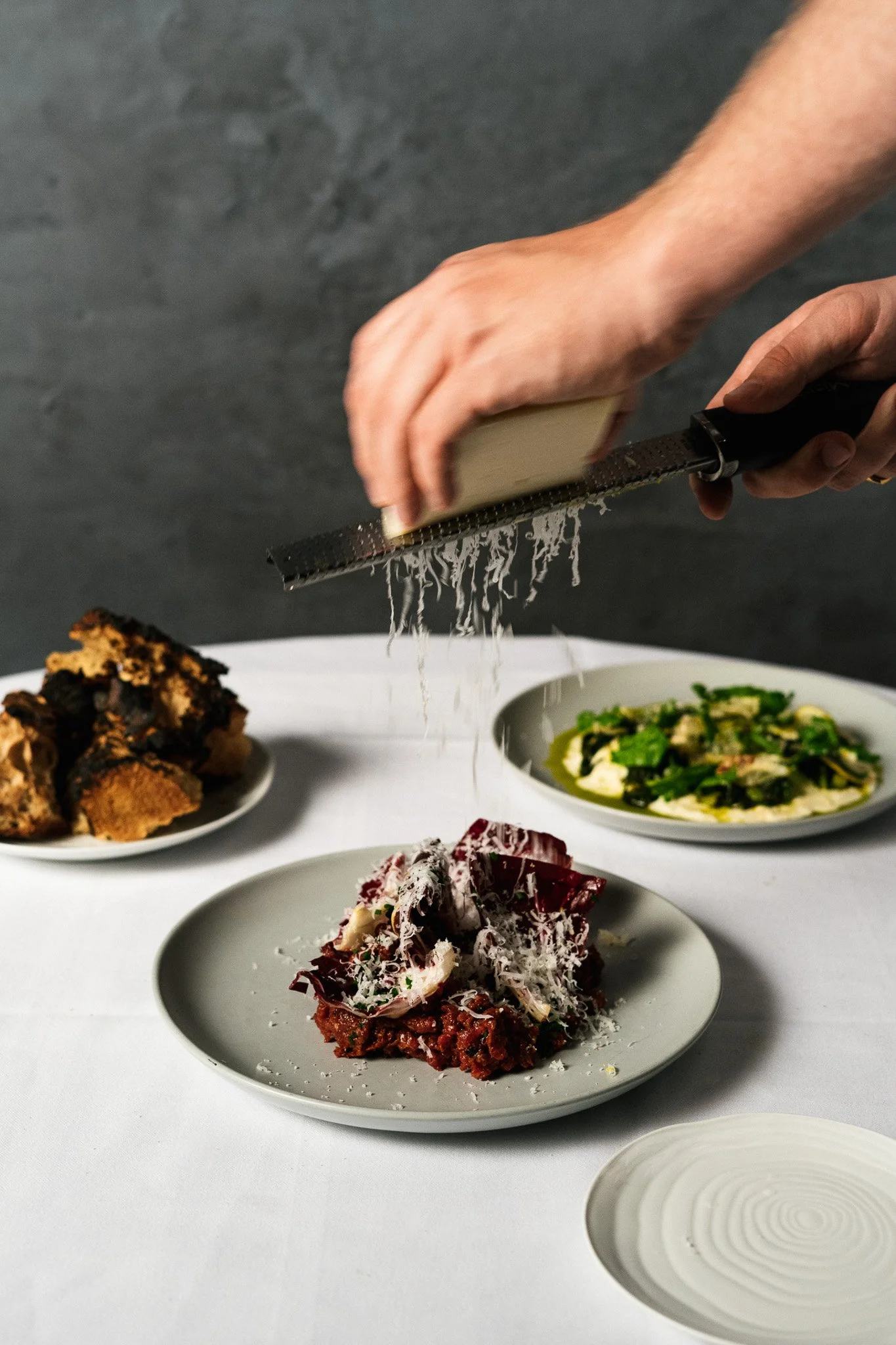 A person grating cheese over a plate of pasta, with other dishes on a white tablecloth in the background.