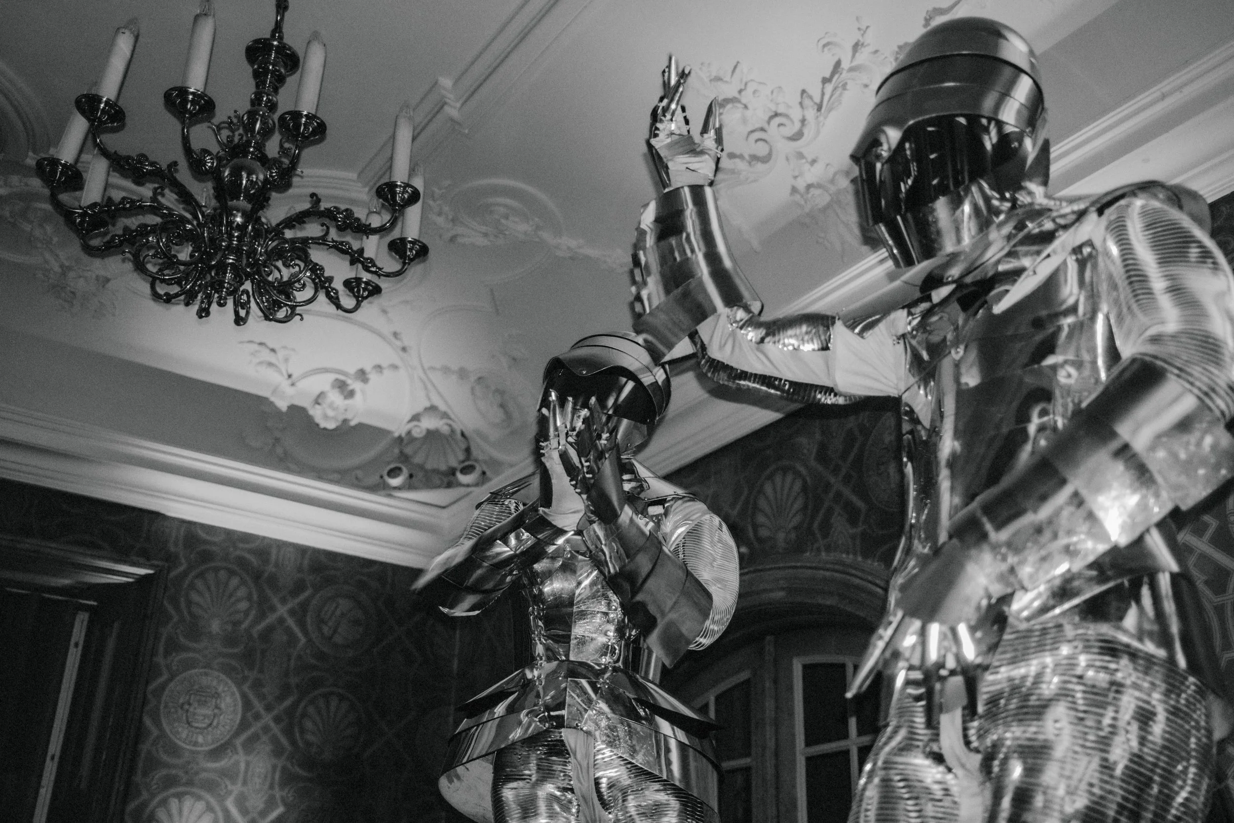 Two people in shiny metallic robot costumes, with one of them covering their face with their hand, standing in an elegant room with a chandelier on the ceiling.