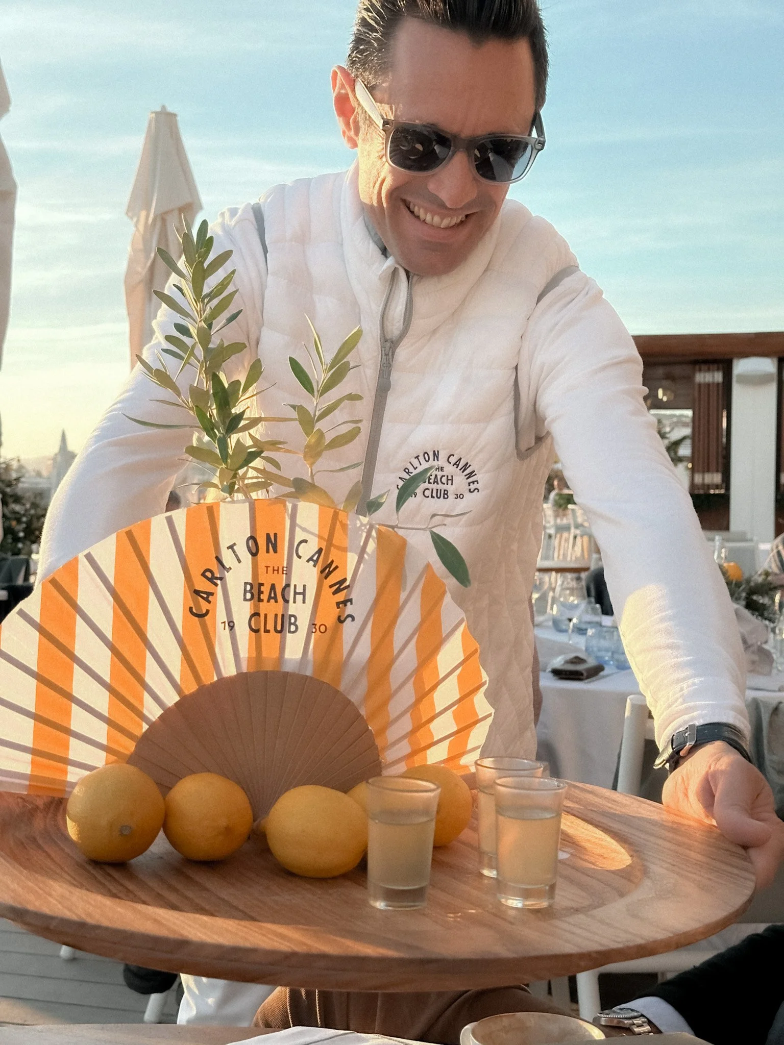 A man wearing sunglasses and a white jacket serving a tray with lemons, a fan, green foliage, and four shot glasses at an outdoor dining area during sunset.