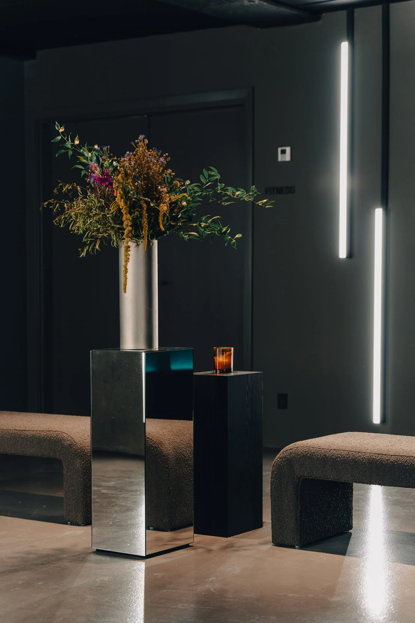 A modern lounge area with a tall flower arrangement in a metallic vase on a reflective metallic stand, two cushioned benches, and minimalistic lighting on dark walls.