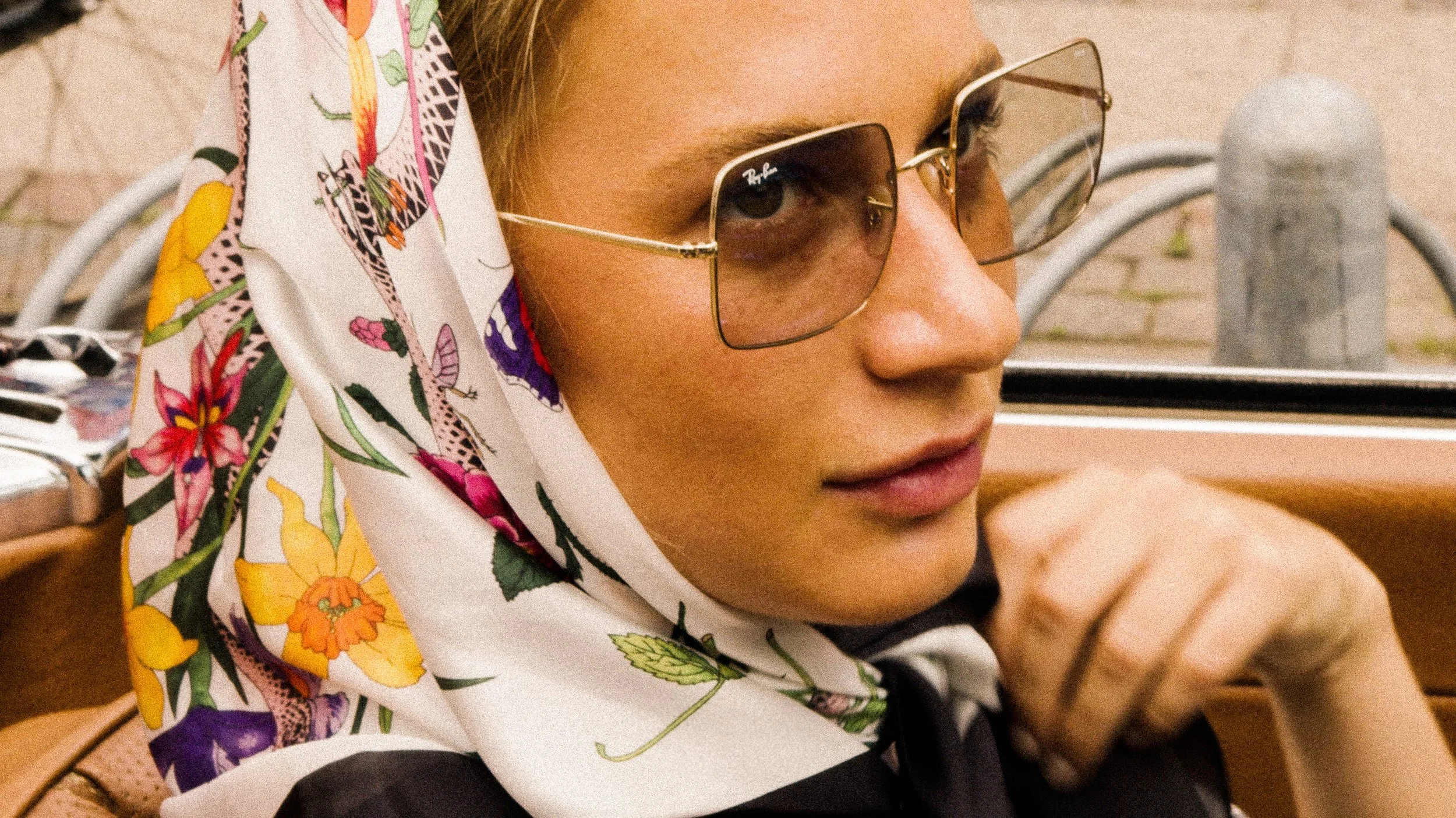 Close-up of a woman wearing glasses and a colorful floral scarf, sitting in a vintage car.