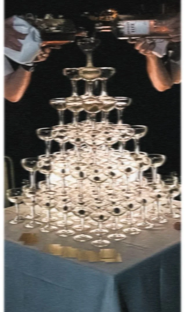 A pyramid of champagne glasses arranged in a tiered formation on a table, with two people in gloves holding bottles pouring champagne into the top glass.