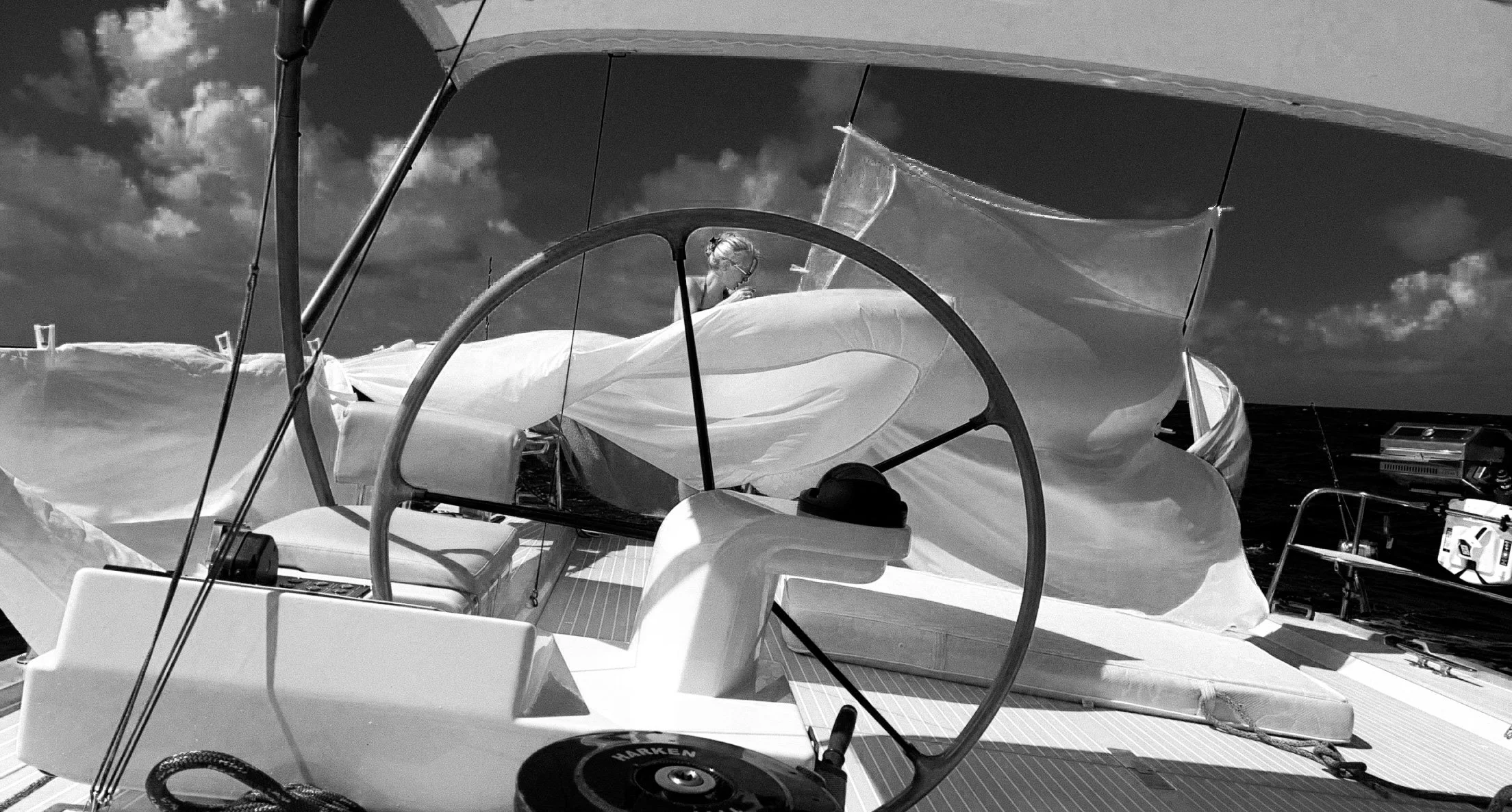 Black and white photo of a woman sitting on a sailboat with its sail partially furled, against a cloudy sky and the ocean in the background.