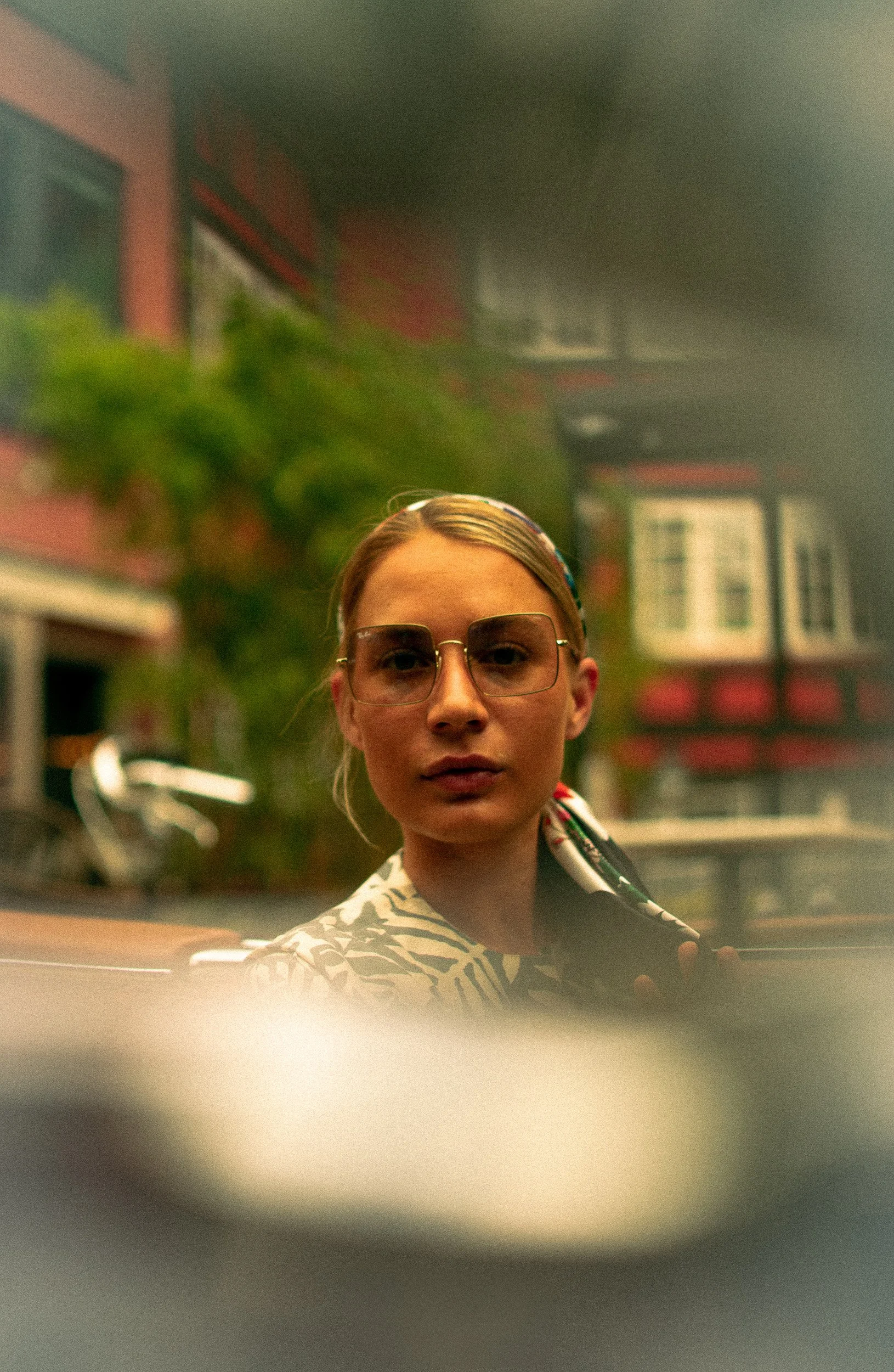 A woman with blonde hair, wearing sunglasses and a patterned scarf, is looking at the camera outside, with a blurred cityscape in the background.