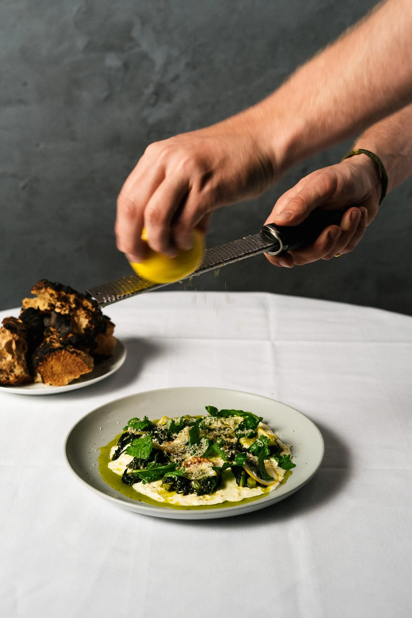 A person grating cheese over a plate of pasta topped with greens, with a plate of bread in the background.