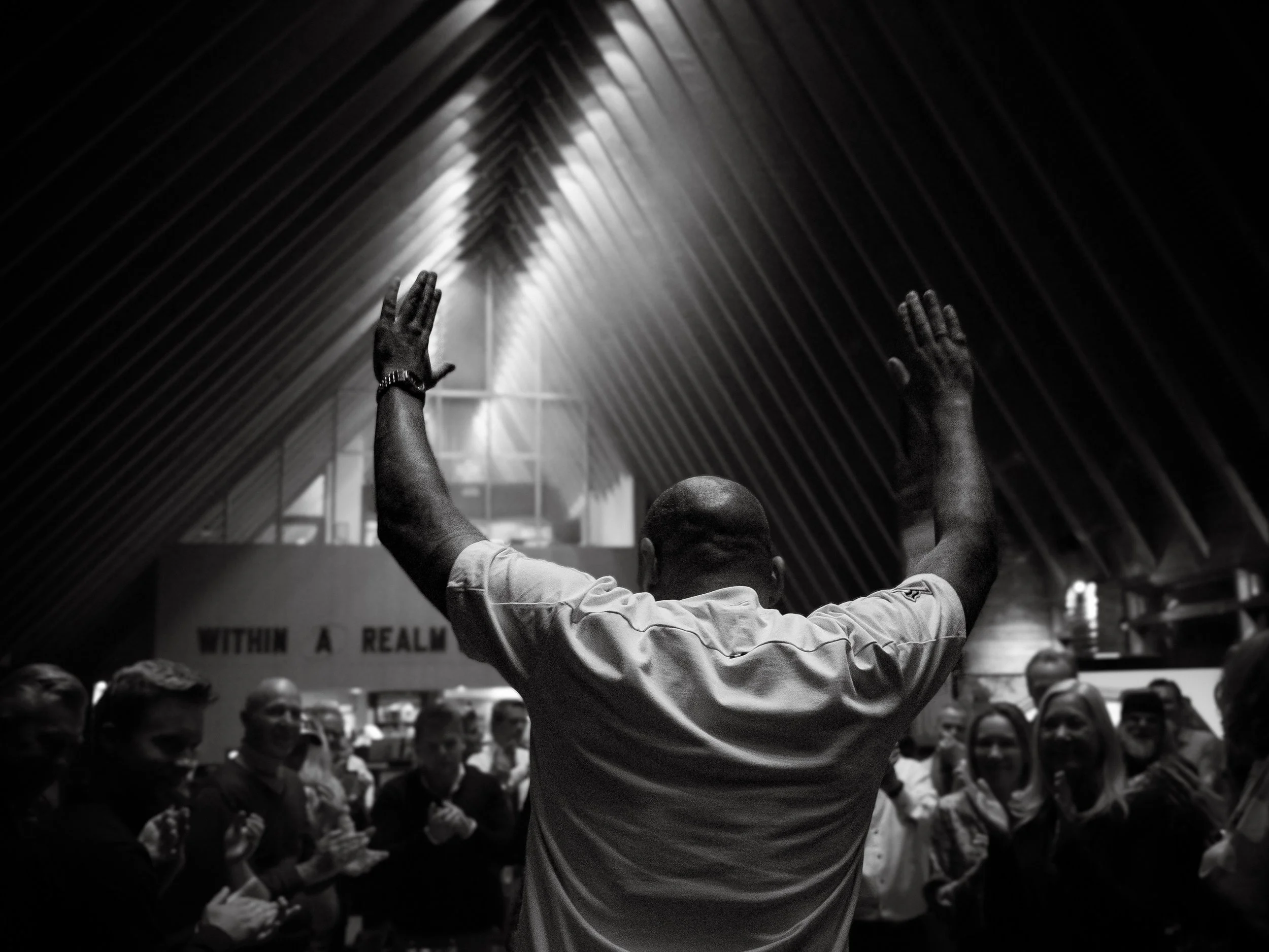 A man with a shaved head and dark skin raising his hands in front of a crowd in a dimly lit indoor space with a high, curved wooden ceiling.