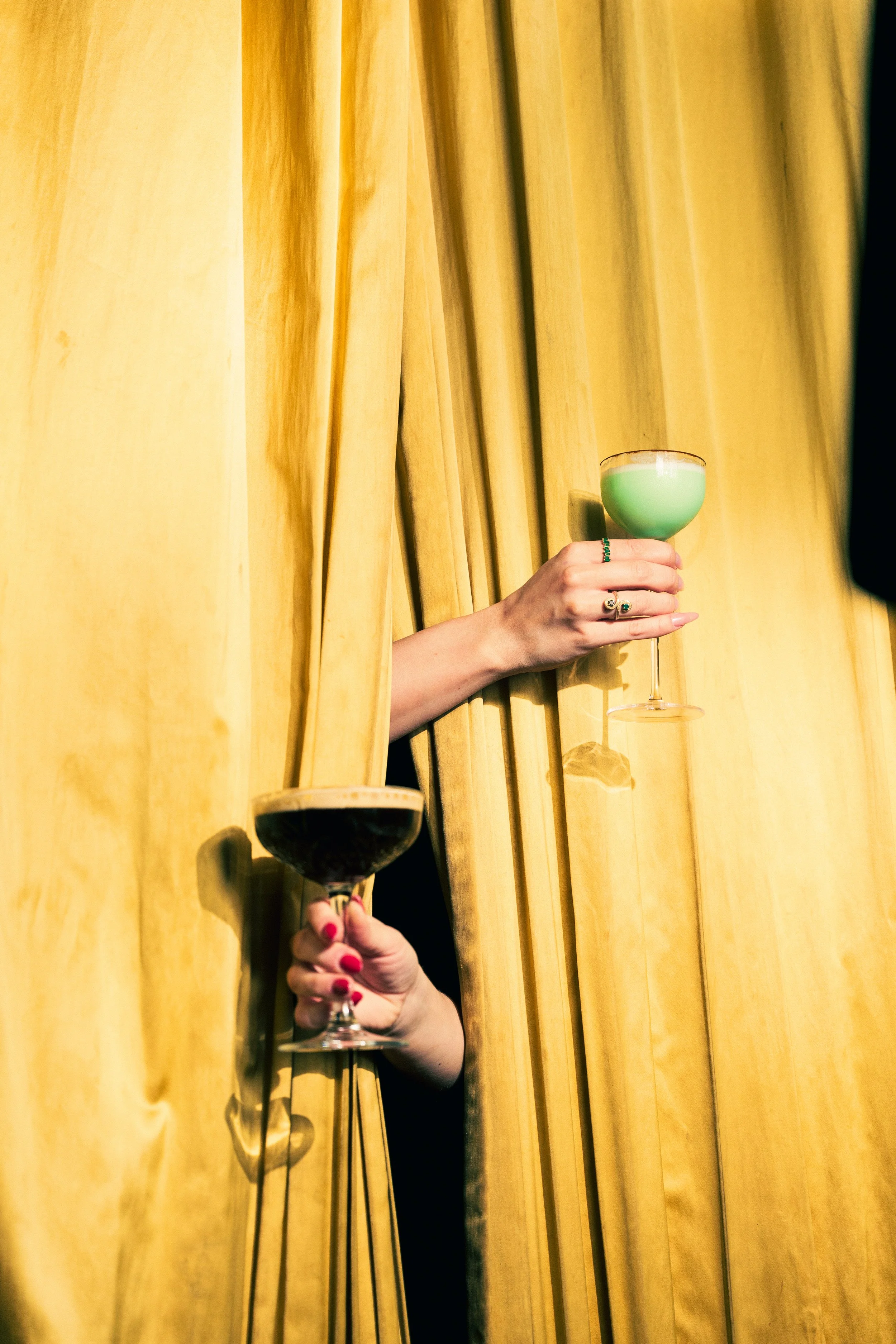 Two hands holding colorful cocktails sticking out from behind yellow curtains. The hand with red nail polish holds a dark-colored drink, and the other hand with green rings holds a green cocktail in a coupe glass.