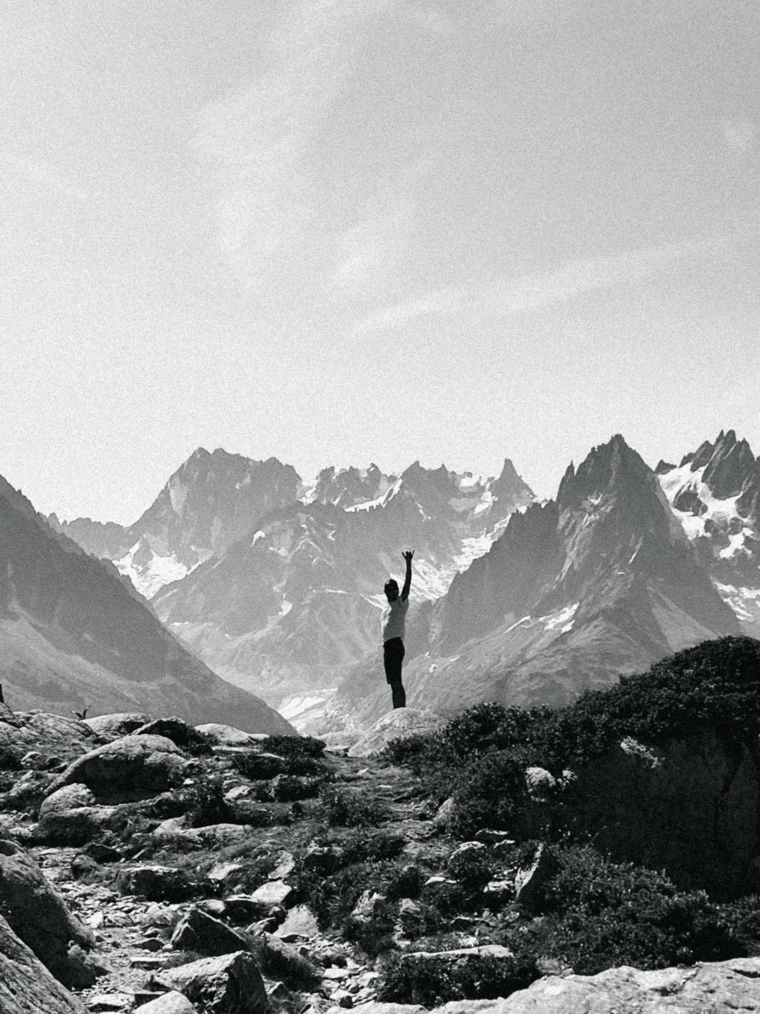 A person standing on rocks in a mountainous landscape with snow-capped peaks in the background, raising one arm in the air.