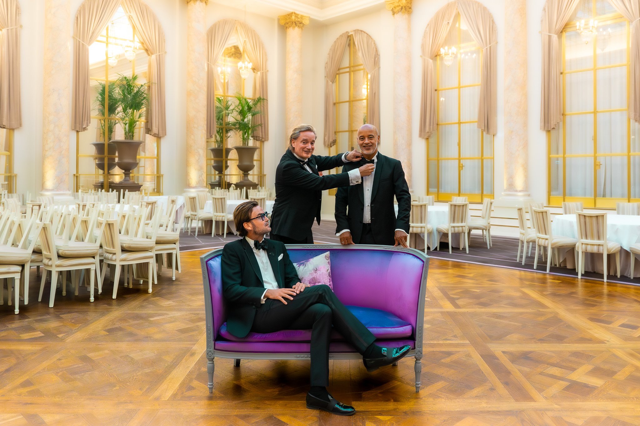 Three men in tuxedos in an elegant ballroom; one seated on a pink and purple couch, one standing behind it adjusting the other's bow tie, and the third standing beside them, smiling.