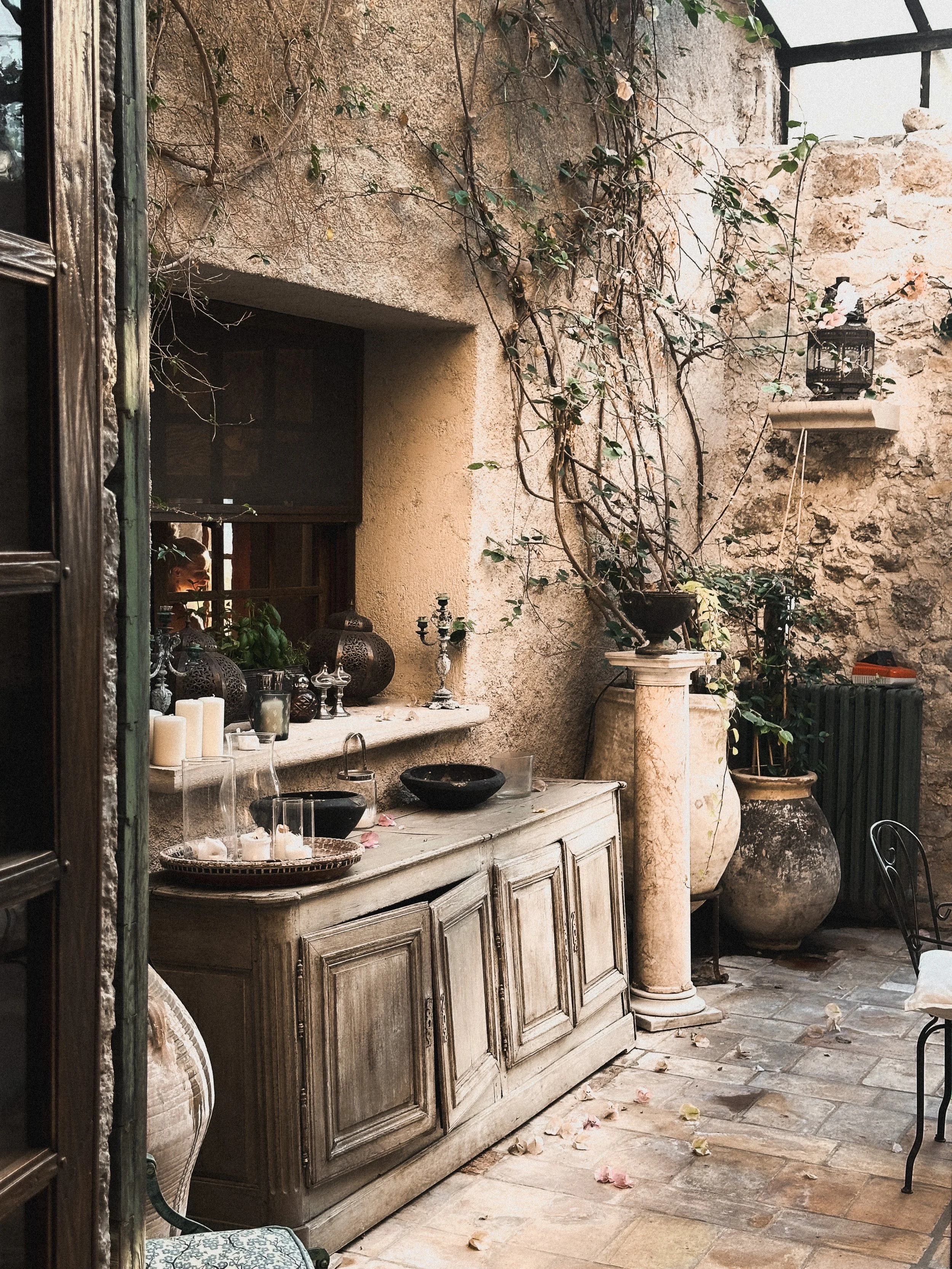 An outdoor stone patio with a vintage wooden cabinet, large decorative vases, candles, and potted plants against a rustic stone wall with vines. There is a window with a person looking inside.