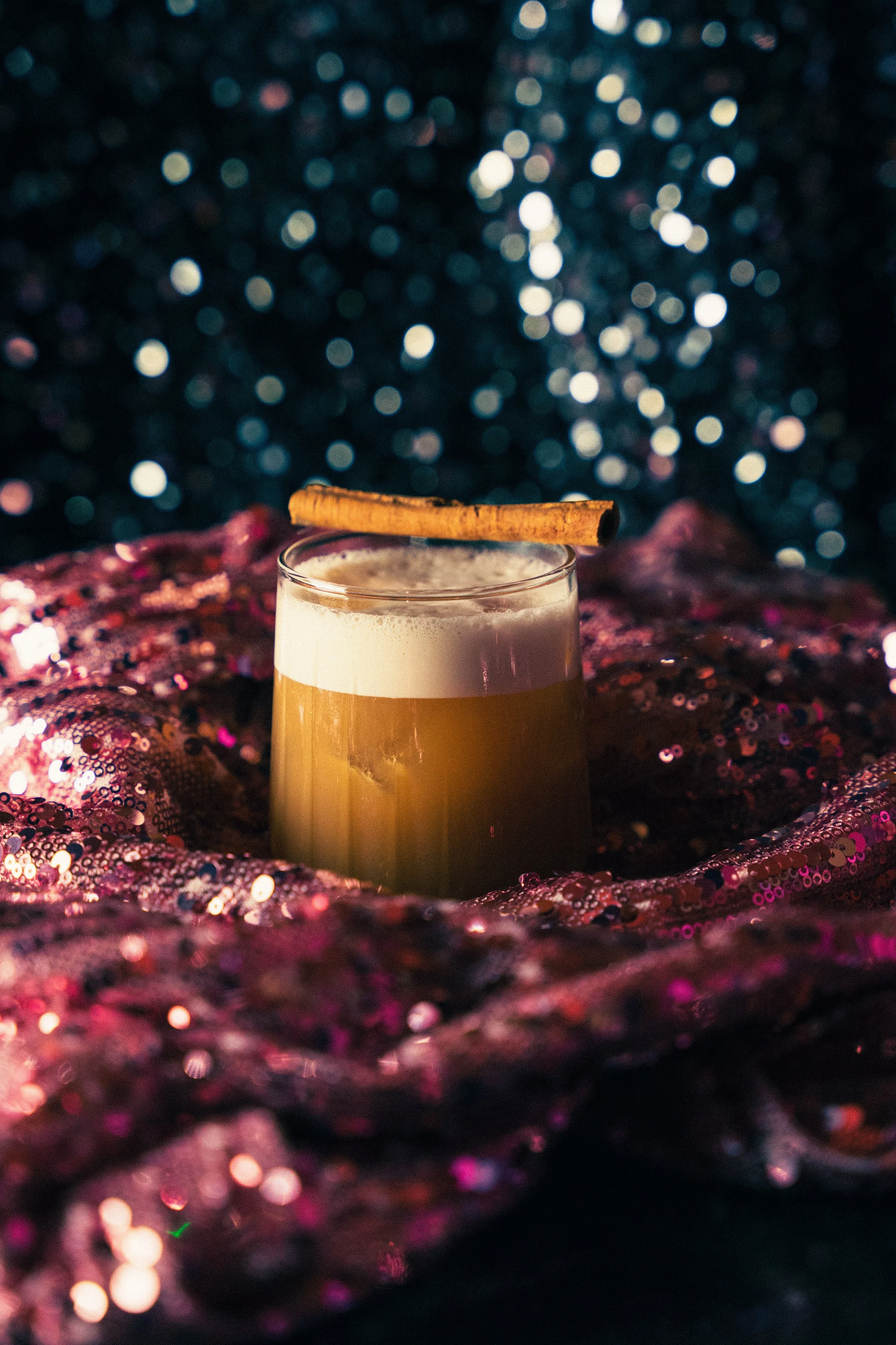 A cocktail glass with a frothy yellowish drink, garnished with a cinnamon stick, sitting on a pink sequined fabric with a dark, bokeh background.